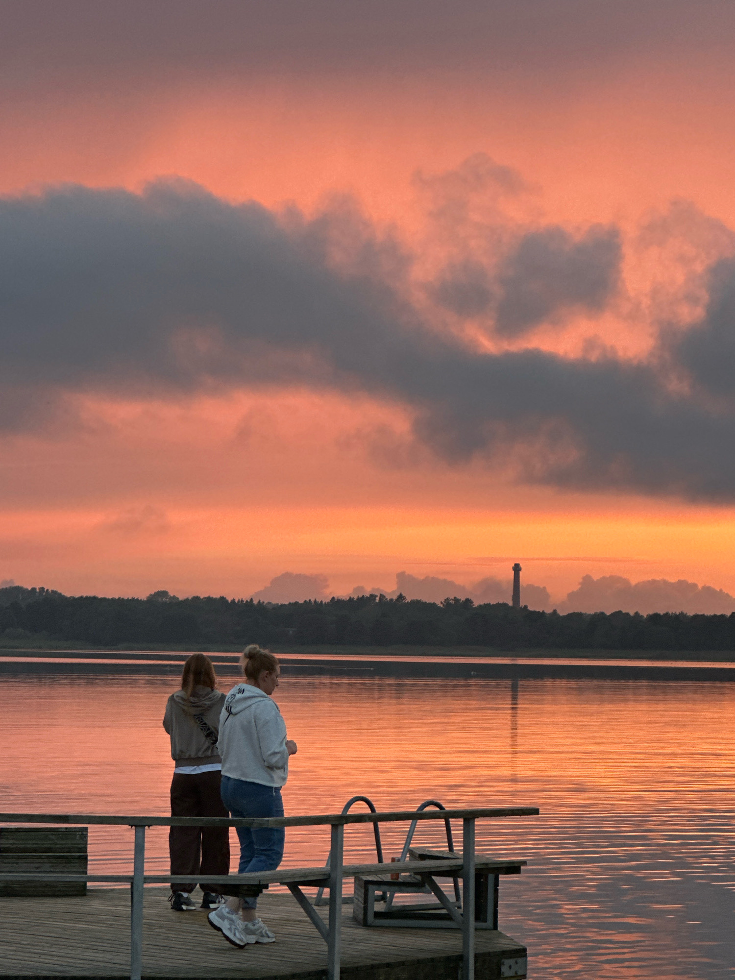 Two individuals stand on a wooden pier overlooking a body of water at dusk. The sky is filled with dramatic clouds painted in shades of orange and pink from the setting sun. A distant lighthouse is visible on the horizon, silhouetted against the colorful sky. The water reflects the sky's vibrant hues, creating a shimmering surface. The pier has railings and a small structure with a bench. The scene is tranquil, with the two people facing away from the camera, seemingly contemplating the view. The location is likely the Kadriorg harbor area in Tallinn, Estonia, known for its picturesque waterfront and views of the Baltic Sea.