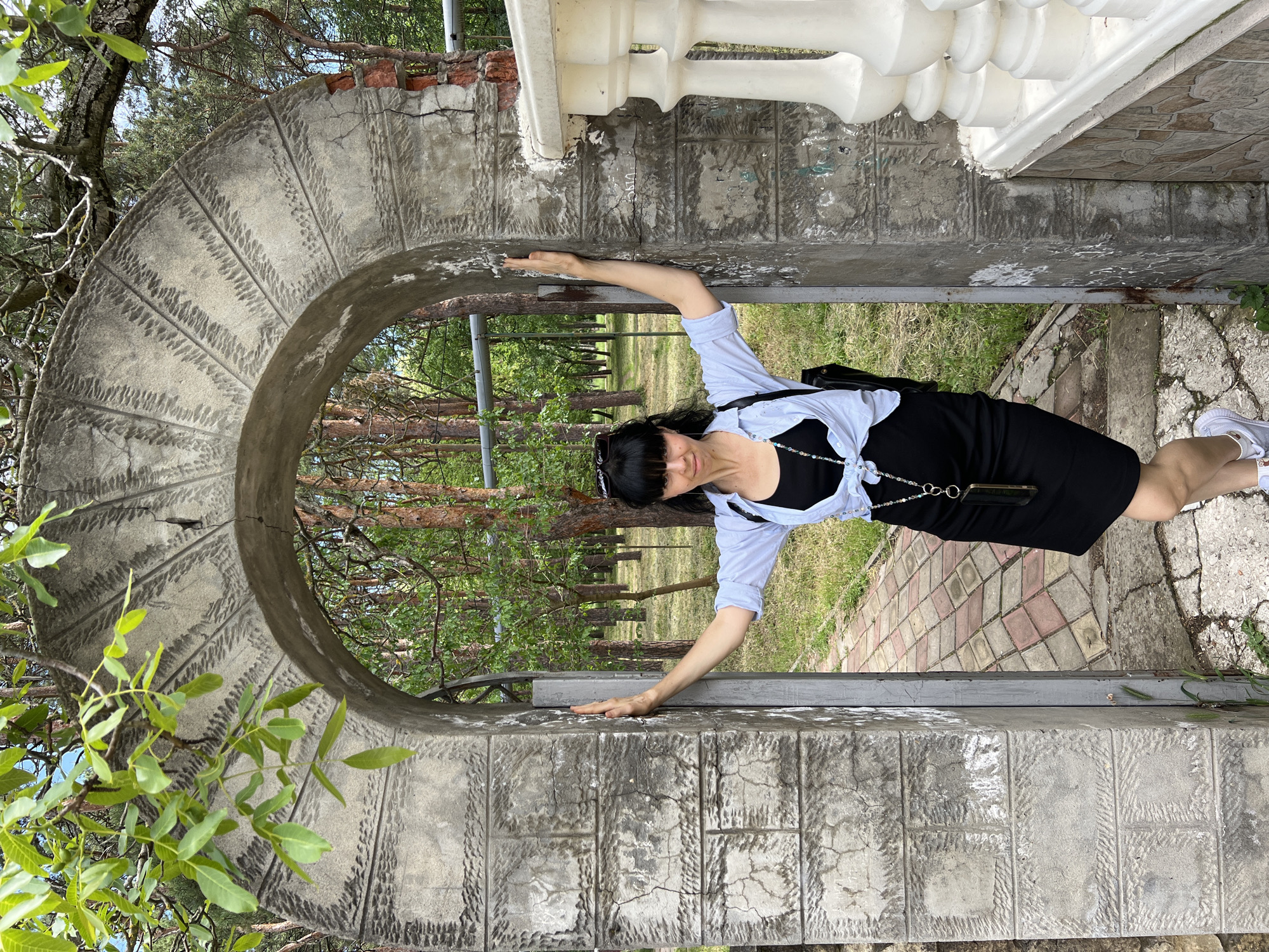 A woman stands within a stone archway, possibly part of an architectural feature, likely within an outdoor space. She has dark hair, is wearing sunglasses atop her head, and is dressed in a black dress with a light blue shirt over it. Her arms are extended, and her hands are placed on the inner edges of the archway. Outside the arch, trees are visible in the distance. The archway appears to be made of gray stone or concrete with a textured surface. The environment suggests a park or garden setting, with vegetation visible around the archway. The location is Krasnodar, Russia.