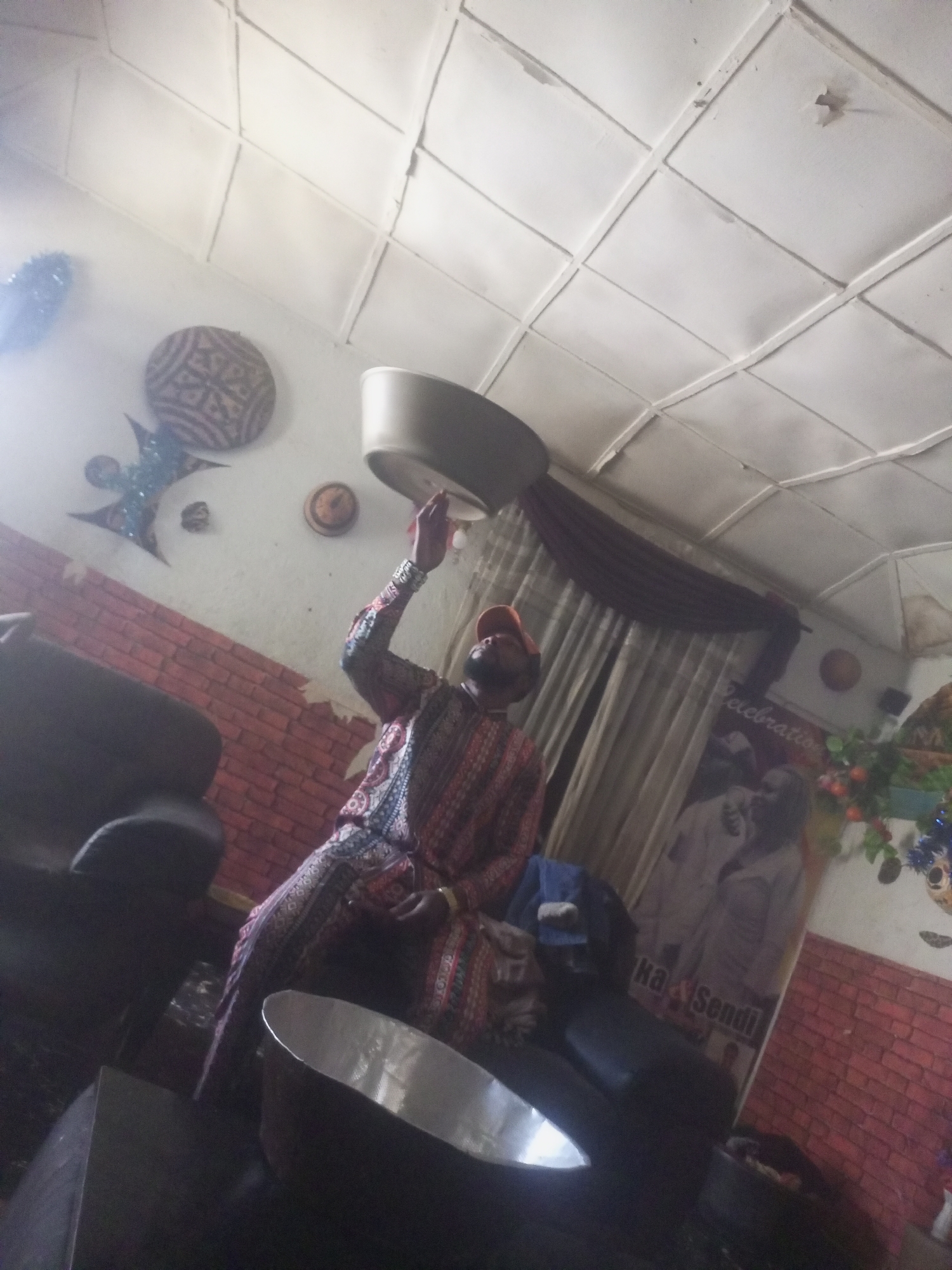 In an indoor setting with a tiled ceiling, a person wearing a vibrantly patterned outfit and an orange cap is seen reaching upwards with their right hand, holding a large, shallow bowl. The person is positioned near a dark armchair. The walls are decorated with a brick pattern in some areas and feature various decorative items, including a circular object with intricate patterns and a dark, star-shaped adornment. In the background, a large poster with text is visible, along with draped curtains and what appears to be a plant with red berries and blue tinsel. A silver-colored basin rests on a surface in the foreground.