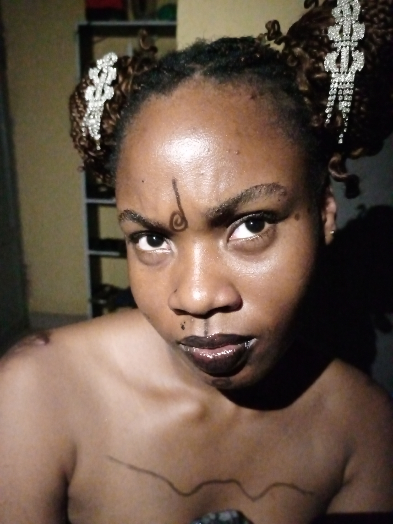 A close-up portrait features a dark-skinned person, likely an adult female, looking intensely and directly into the camera. Her hair is styled in two prominent high buns, each secured with a sparkling silver, rhinestone-studded hair clip shaped like a dollar sign ($). A distinctive, stylized dark spiral design is drawn vertically on her forehead between her eyebrows. She wears dark, glossy lipstick, and a small black stud nose ring pierces her left nostril. Her eyebrows are furrowed, contributing to a serious or defiant expression. Her shoulders and upper chest are bare, revealing a thin, dark line drawn across her décolletage, possibly as body art. The background is an indoor setting with a blurred, dark shelf unit partially visible behind her left shoulder, illuminated by artificial light that creates highlights and shadows on her skin.
