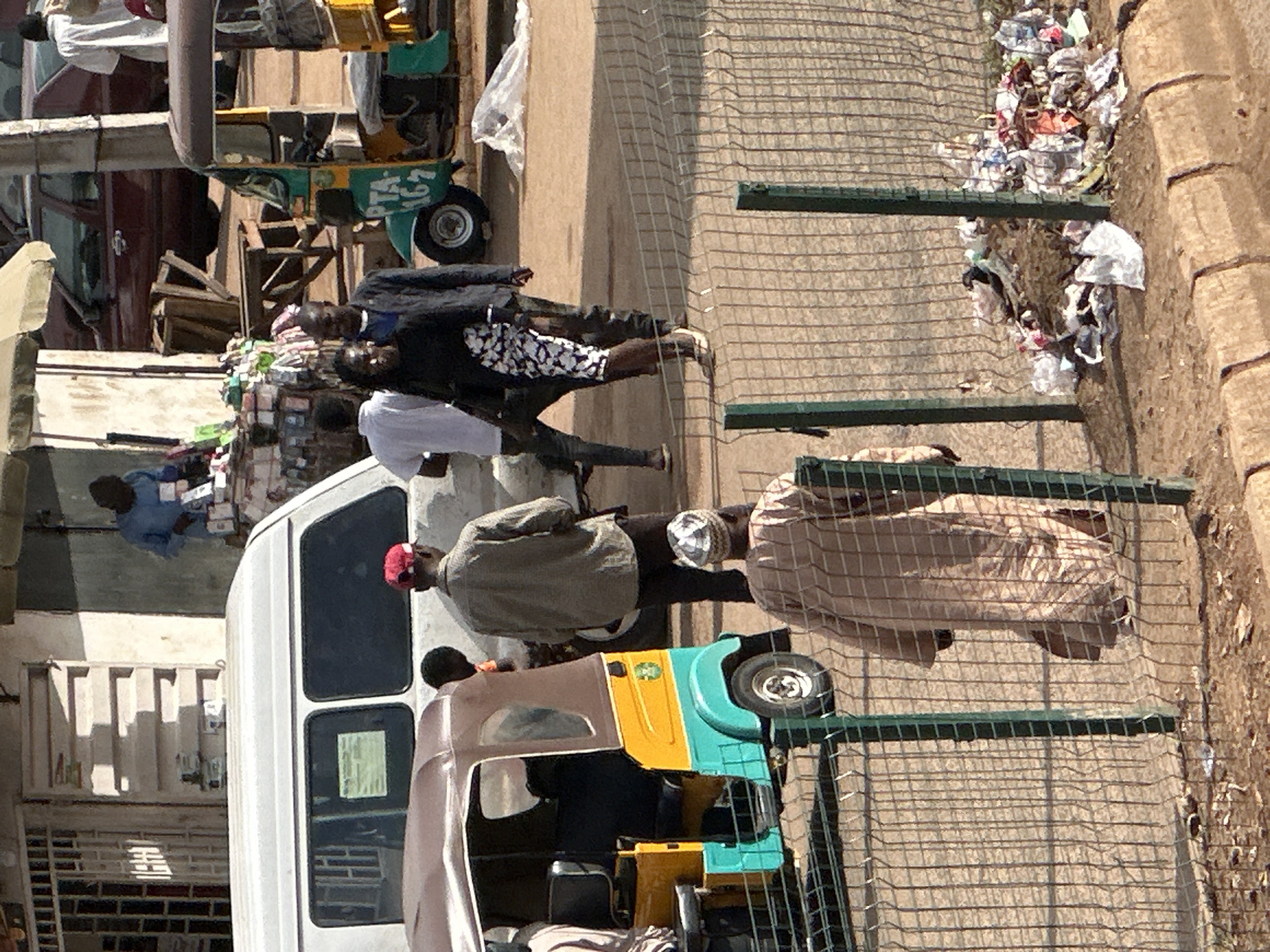 This image depicts a bustling street scene in Bukuru, Nigeria, with a focus on human activity and modes of transportation. In the foreground, a fence separates the viewer from a pile of refuse. Beyond the fence, a colorful auto-rickshaw, painted yellow, turquoise, and brown, is parked. Two individuals are visible near the auto-rickshaw; one is seated inside, and another, wearing a patterned headscarf and a tan robe, walks away from the vehicle.
Behind the auto-rickshaw, a white van is parked. Several people are in the vicinity of the van and a market stall. A man in a grey shirt and red cap stands by the van, facing away from the viewer. To his left, a man in a white shirt and dark pants is also visible. Further to the left, a couple walks together, with the woman wearing a black top and a patterned skirt.
In the background, the market stall features an array of goods, including packaged items and what appear to be medicines or toiletries displayed on shelves. A man in a blue shirt is attending to the stall. A maroon car is parked on the street, and a person in a white garment stands near it. The environment appears to be a dusty, open area with buildings and market structures. The overall impression is one of everyday life and commerce in a Nigerian urban setting.
