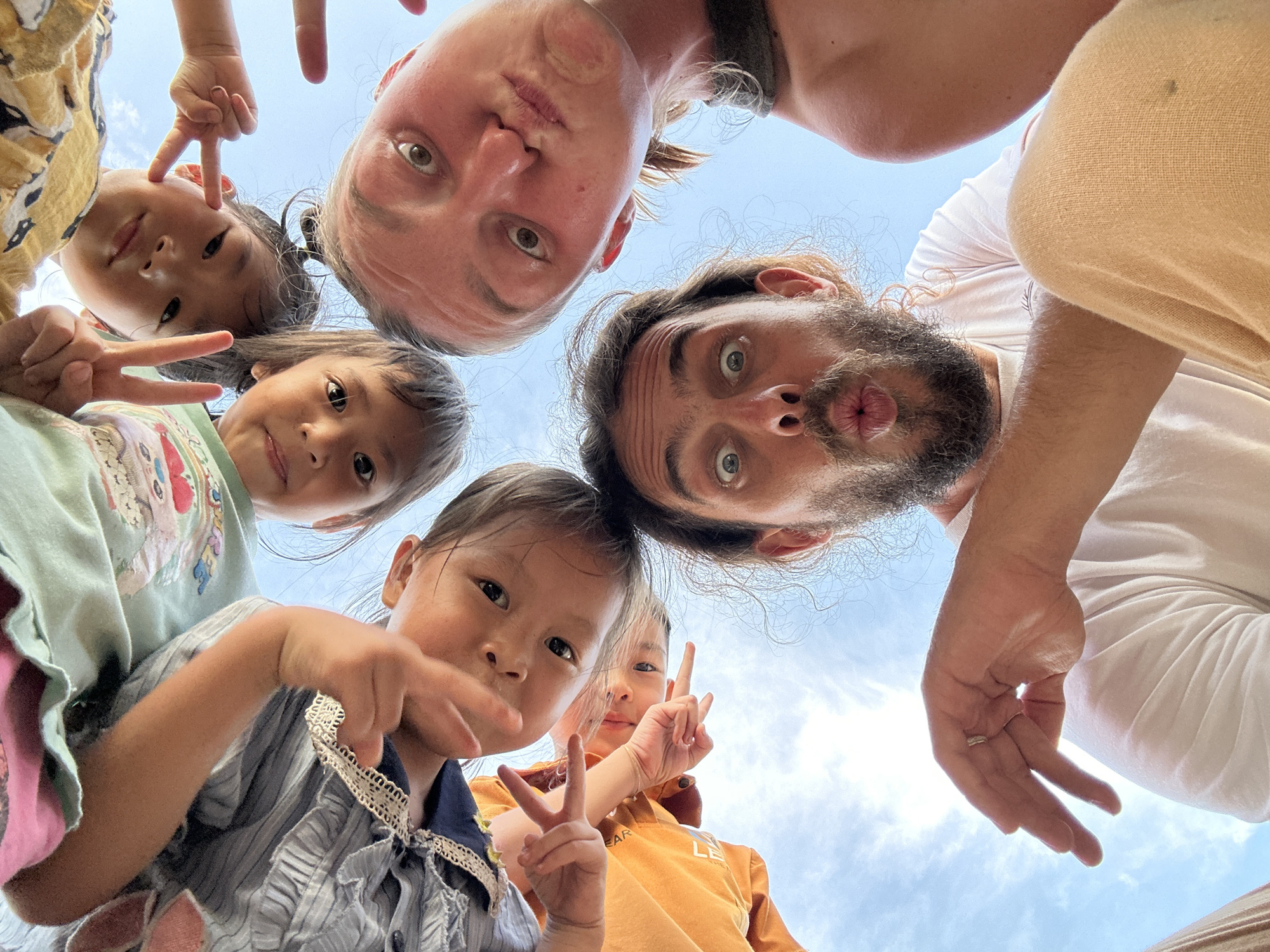 A low-angle, upward-facing photograph captures seven individuals – two adults and five children – gathered in a circular formation, leaning over the camera. The background is a clear, bright blue sky with sparse white clouds, indicating an outdoor, daytime setting.
In the bottom center, a male adult with a full dark beard, long dark hair, and light skin gazes directly into the camera with wide eyes and puckered lips. He wears a white t-shirt, and his light beige pants are visible at the bottom right. A silver ring is present on his left ring finger. To his right, an adult female with light skin and light brown hair displays a similar wide-eyed, puckered-lip expression. She wears a dark olive-green sleeveless top.
Five children surround the adults at the top of the frame. One child wears a light green t-shirt with a graphic design and holds up a peace sign with their left hand. Another child, wearing a yellow patterned shirt, also makes a peace sign with their right hand. A third child, in a blue and white striped dress with white lace trim, has their left hand extended towards the camera. A fourth child, in an orange t-shirt, also shows a peace sign with their right hand. A fifth child, partially visible behind the child in the blue dress and orange shirt, also looks down. The children exhibit varying expressions, predominantly playful or neutral, and many are looking directly into the camera.
The scene is located in Gia Hội, Vietnam.