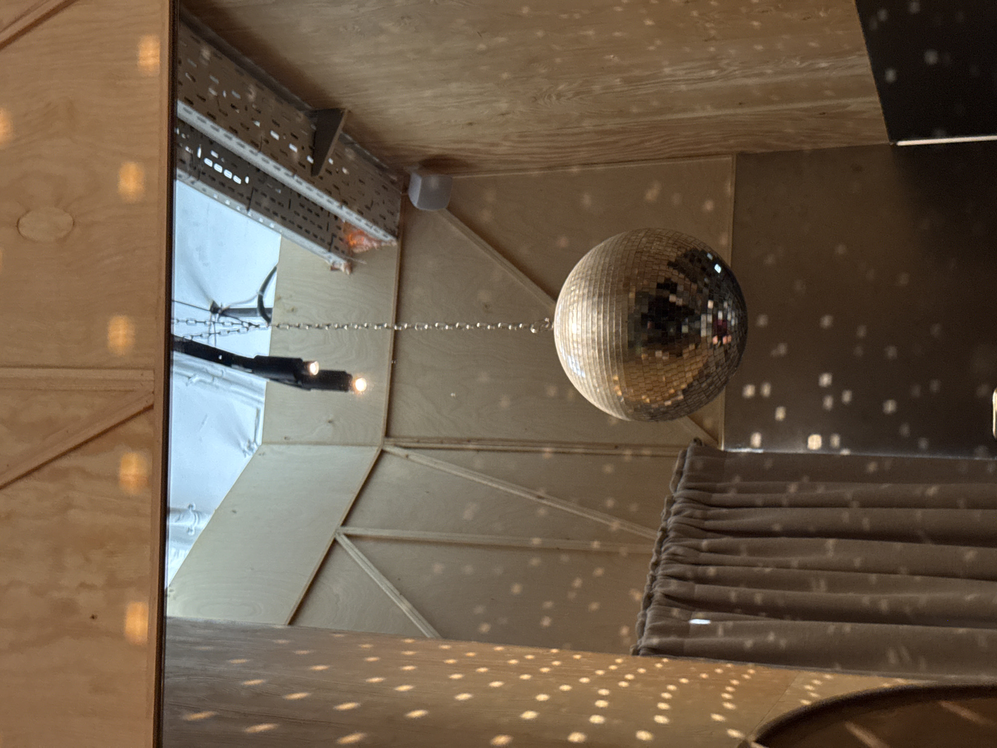 The image shows an indoor space with wooden walls and ceiling. A disco ball is hanging from the ceiling by a chain in the center of the frame. There are lights reflecting off the disco ball and onto the walls, creating a speckled pattern. A curtain hangs on the left side of the image. In the background, there is a metal structure with wires attached to it, and a speaker mounted on the wall. The location appears to be a bar or club in Estosadok, Russia. There are no people visible in the image.