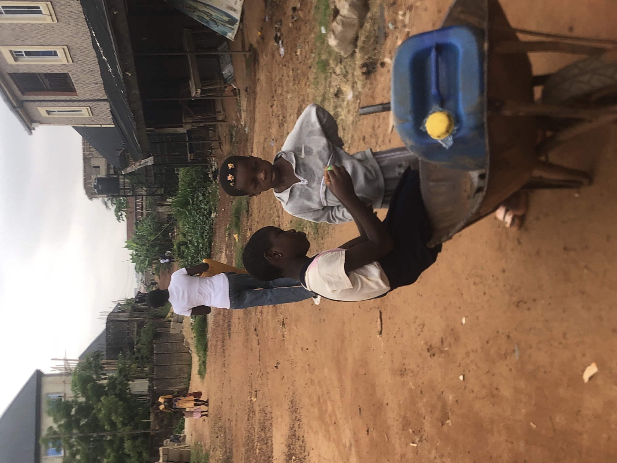 The image depicts an outdoor scene in Ukwu-Abwa, Nigeria, characterized by unpaved, dusty ground and a mix of residential buildings and vegetation under an overcast sky.
In the immediate foreground, two children are the central subjects. One child, wearing a grey Nike-branded hoodie and with small decorative elements in their hair, stands facing another child. This second child, seen in profile, wears a white and dark-colored top and is looking up at the first child, suggesting an interaction or conversation. To their right, a rustic metal wheelbarrow holds a blue plastic jerrycan with a distinctive yellow cap.
Further back on the left side, a man wearing a white t-shirt and jeans is visible, appearing to walk or stand while holding a yellow container. In the distant background, two smaller figures, possibly children, are partially obscured.
The setting includes various buildings. To the left, a finished two-story building with a textured grey facade and white-framed windows is prominent. Other structures, some appearing to be under construction with visible scaffolding, are scattered across the background. Patches of green vegetation, including bushes and trees, are interspersed throughout the scene. The overall impression is a glimpse into daily life in a developing community. The visible "Nike" logo on the hoodie is a notable detail.