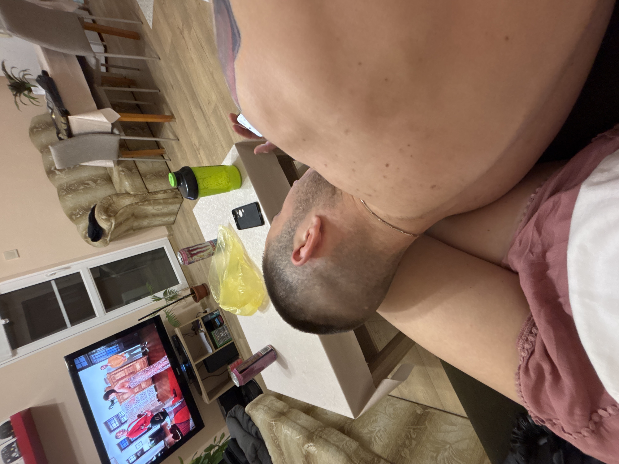 Here is a forensic description of the image:
**Scene:** Interior of a residential space, potentially an apartment or house. The image is taken from a high angle, looking down onto a table and a person.
**People:** Two individuals are partially visible.
* Person 1: Appears to be lying down or reclining on a surface. Part of their back, shoulder, and upper arm are in view. A tattoo is visible on their back.
* Person 2: Only the head is visible. This person has short, cropped hair and a gold chain around their neck.
**Objects:**
* Table: A white, rectangular table is the central point of the foreground.
* Electronic Devices: A smartphone is on the table.
* Beverages: A green beverage bottle and a can are present on the table.
* Groceries: A plastic shopping bag filled with food.
* Furniture: Chairs, a couch, and a shelving unit or entertainment center.
* Television: A television is visible, with a program playing.
* Plants: A potted plant is near the shelving unit.
* Pillows: Pillows are on the couch
**Actions/Interactions:** It is impossible to determine what activities are taking place, besides the TV is playing a show.
**Environment:** The room seems to be a living area or a combination of living and dining spaces. Lighting is a combination of natural light from the window and possibly artificial light.
**Location:** Unknown, with no landmarks or street signs to identify the location in Unknown, Unknown.