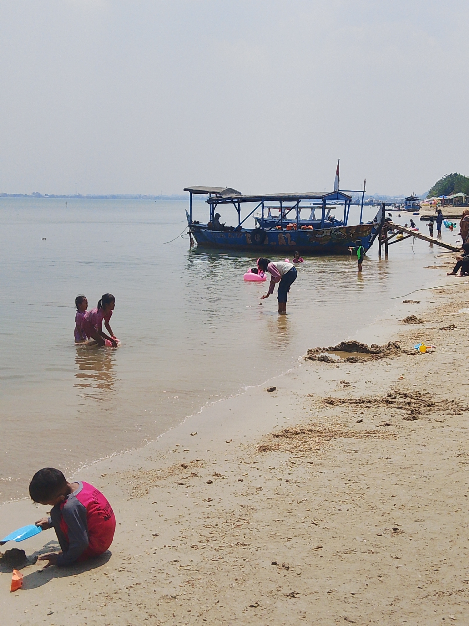 The image depicts a sunny day at a beach in Pejagan, Indonesia, with several people engaged in various activities. In the foreground, a young boy is sitting on the sand, playing with a blue plastic shovel and some dirt. Further out in the shallow water, two young girls in pink swimwear are playing together. A larger blue boat with a canopy is moored offshore, with people visible on and around it, and another smaller boat further in the distance. The beach has a sandy shore with some scattered debris, and the water is calm and appears shallow. In the background, a distant shoreline with vegetation and structures is visible under a hazy sky. The overall scene conveys a casual, family-friendly atmosphere at a coastal recreational area.