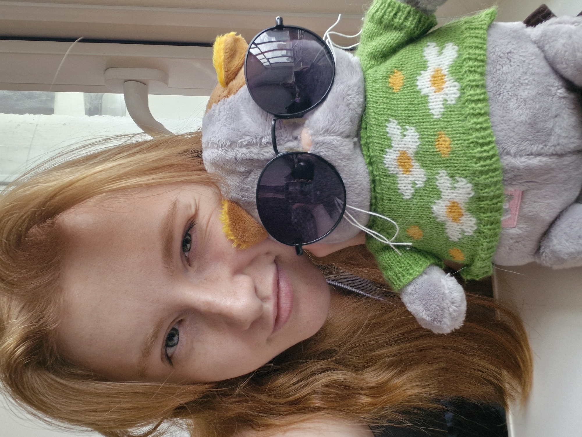 A selfie shows a fair-skinned woman with light red hair, light eyebrows, and blue eyes, rotated sideways. She is holding a stuffed animal against her head. The animal is gray with yellow ears and wears sunglasses and a green sweater with white daisies. Reflected in the sunglasses are a building and a cloudy sky. The window frame behind the woman is white and shows light reflecting on it, and a short line of white snow can be seen outside of the window. This scene is unlikely to be in Yekaterinburg, Russia, as it has a continental climate with predominantly cold winters.