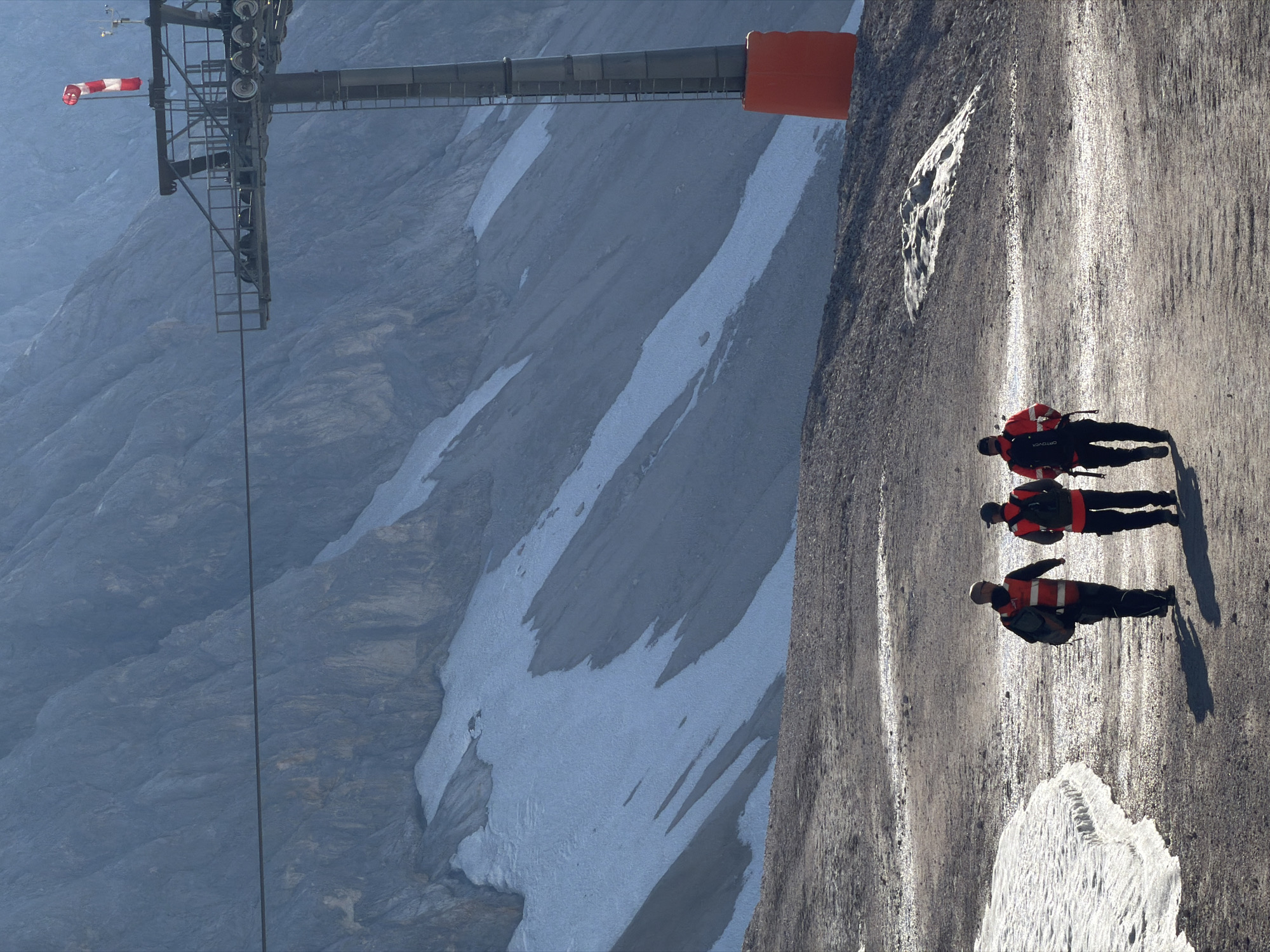The image depicts three individuals wearing high-visibility vests and backpacks walking along a gravel path on a mountainside. The background features a large, rocky mountain slope with patches of snow. In the upper right portion of the image, a ski lift pylon with a red safety bumper and a windsock is visible, indicating an alpine environment. The overall scene suggests a location in a mountainous region, likely in the vicinity of Garmisch-Partenkirchen, Germany, known for its alpine tourism and ski resorts. The people appear to be engaged in some form of outdoor activity or work, possibly related to the ski resort.