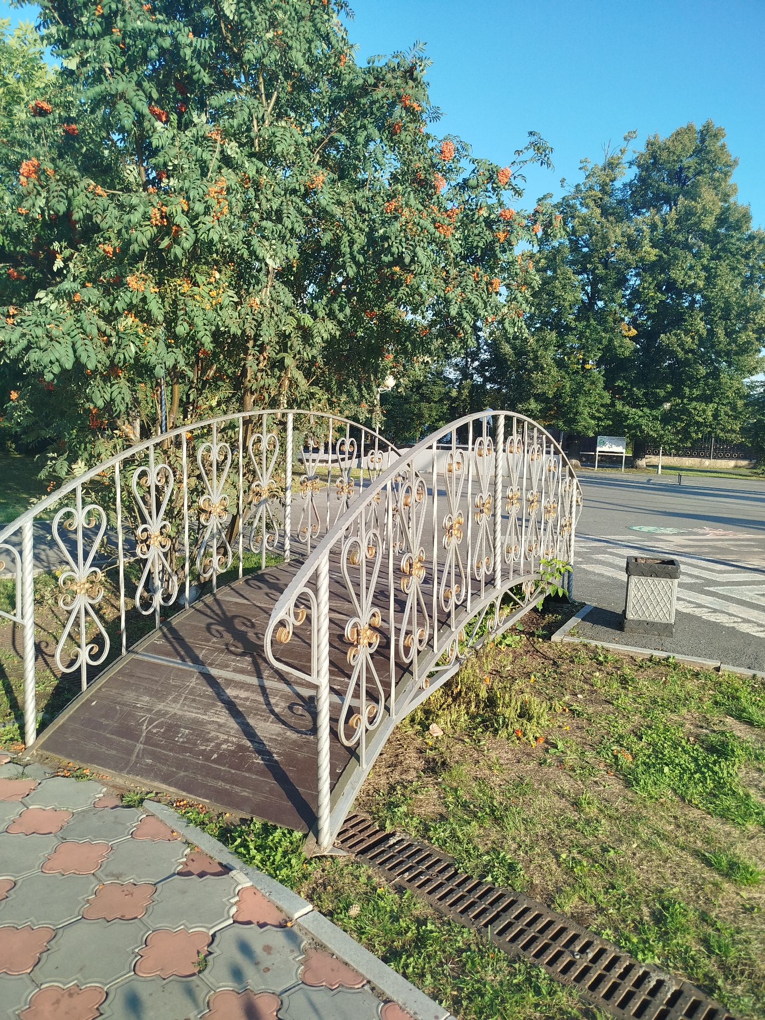 The image shows a decorative metal bridge with intricate scrollwork, spanning a small grassy area. The bridge has wooden planks for the walkway and a white metal railing adorned with gold accents. The bridge is situated next to a paved area with patterned tiles and a road beyond. A metal trash receptacle with a diamond pattern is visible near the road. In the background, lush green trees, one laden with bright orange berries, and a clear blue sky are present. The scene appears to be in an outdoor urban park or recreational area. The trash receptacle has a sign that indicates it's located in Yalutorovsk, Russia.
