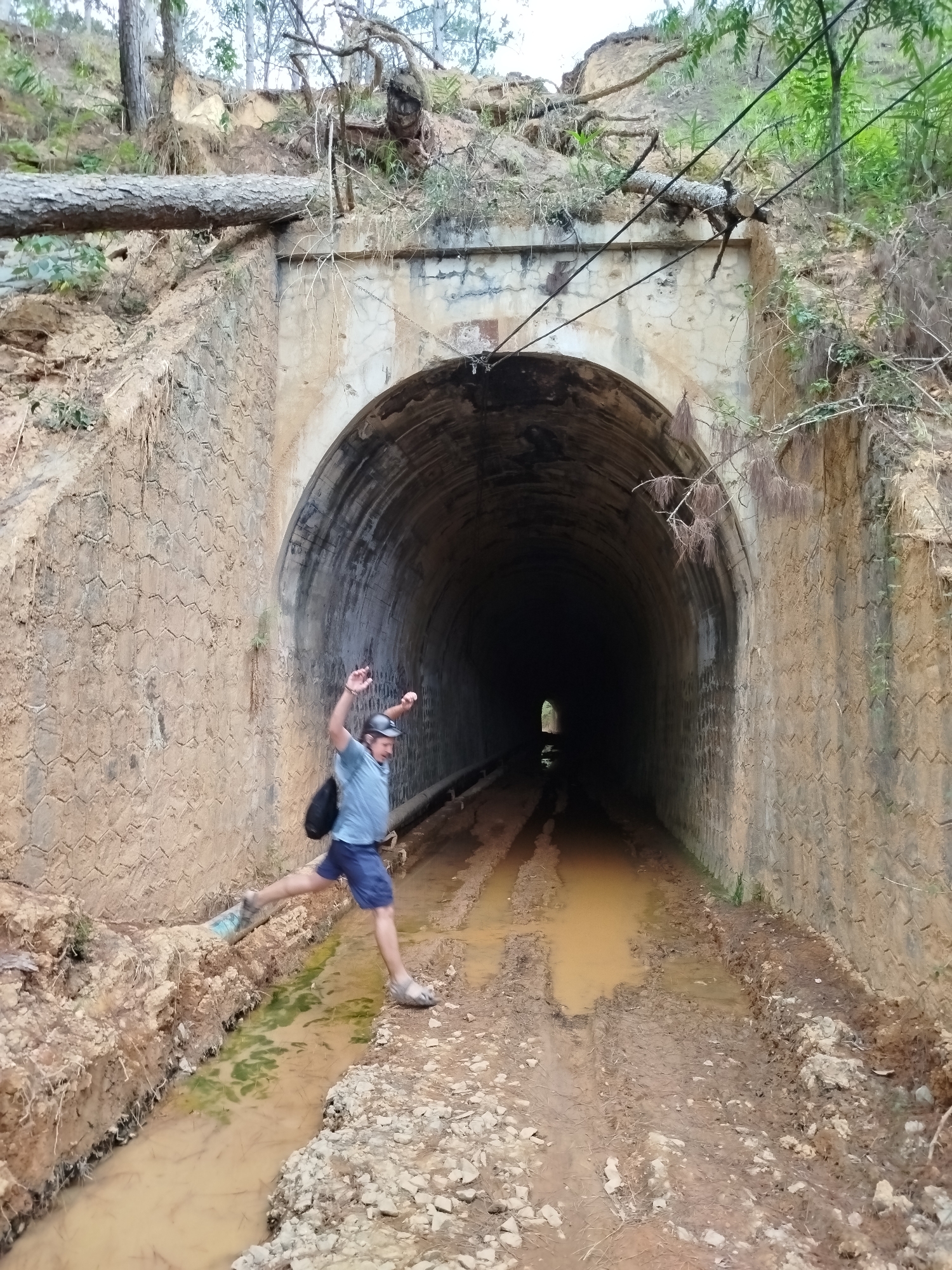 A man in a blue shirt, navy shorts, and a baseball cap is jumping across a muddy stream in front of a bricked tunnel. The tunnel is located in Đa Thọ, Vietnam. The tunnel opening is arched and appears to be crumbling. A fallen tree lies across the top of the tunnel entrance. Electrical lines are strung above the tunnel opening. The tunnel interior is dark and muddy. The foreground is muddy with a flowing stream. The surrounding vegetation is sparse.