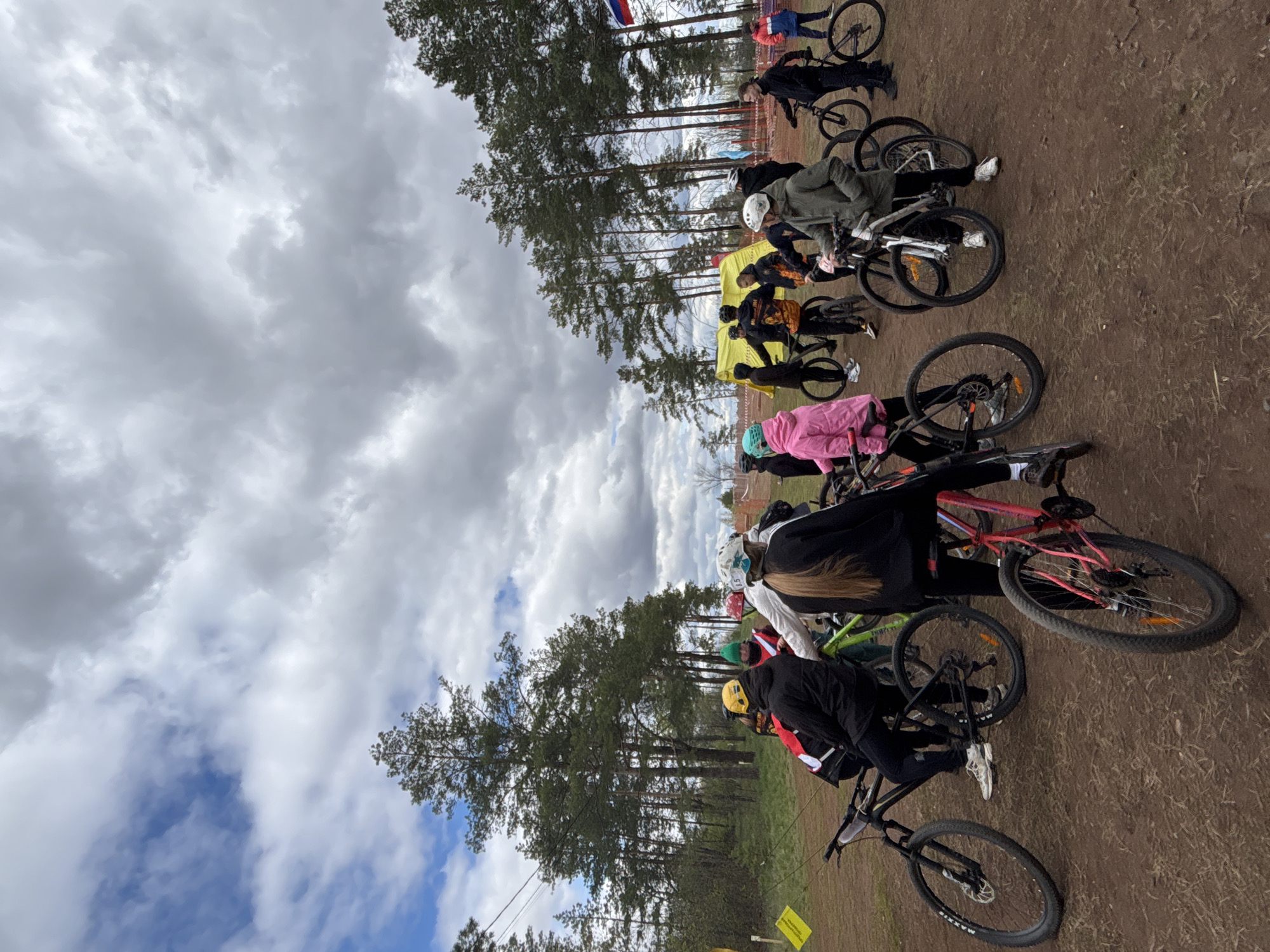 A group of people on bicycles are gathered on a dirt path next to a line of pine trees. The sky is cloudy with patches of blue. Some individuals are wearing helmets and brightly colored jackets. A yellow tent is visible in the background. A Russian flag is present. The location is outdoors, likely in or near Orenburg, Russia.