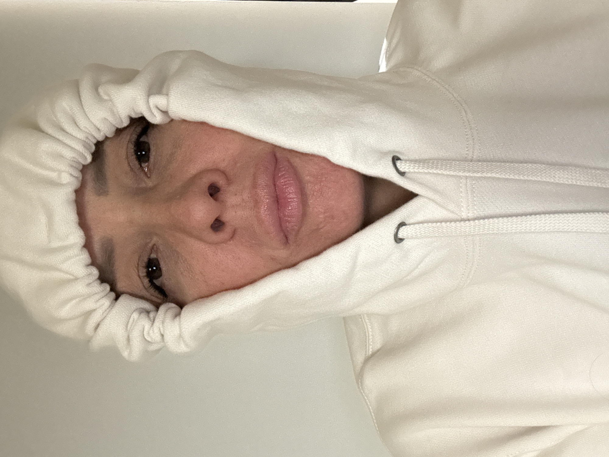 A close-up, slightly rotated image shows a person wearing a white hooded sweatshirt. The hood is up, partially obscuring the face, with only the eyes, nose, and mouth visible. The person's skin appears light-toned, and the lighting is soft and even. The background is a plain white surface. There is no indication of the location being Chaikovsky, Russia, or any other specific city.