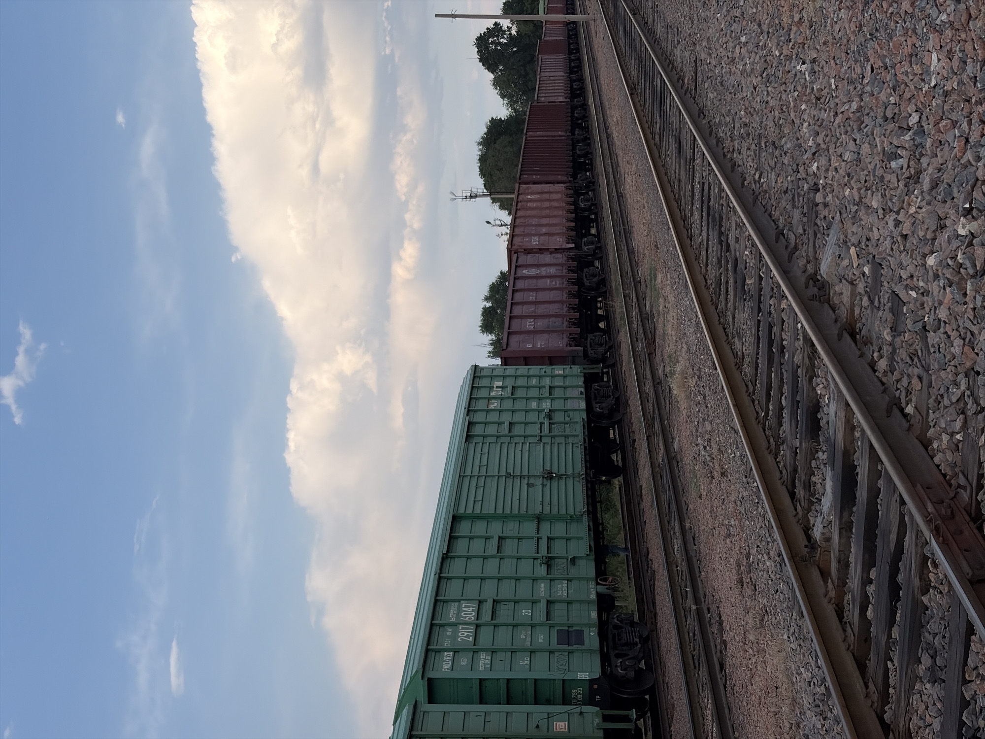 The image shows a freight train on a railway track under a partly cloudy sky. The train consists of several boxcars, some of which are dark red and others are a vibrant green. The tracks are surrounded by gravel and some dry grass. Trees are visible in the background, suggesting a rural or semi-rural setting. The time of day appears to be late afternoon or early evening, given the quality of light. There are no people visible in the image.
Notable details include the license plate or identification numbers on the green boxcar, which appear to read "2917 6047" and "10799". Other markings on the green car include "PKD/RZD" and what seem to be weight specifications. The dark red boxcars also have markings, but they are less legible. The presence of railway infrastructure suggests this is a functional transportation route. The overall mood is one of stillness and industrial activity in a natural landscape. The location is identified as Kant, Kyrgyzstan.