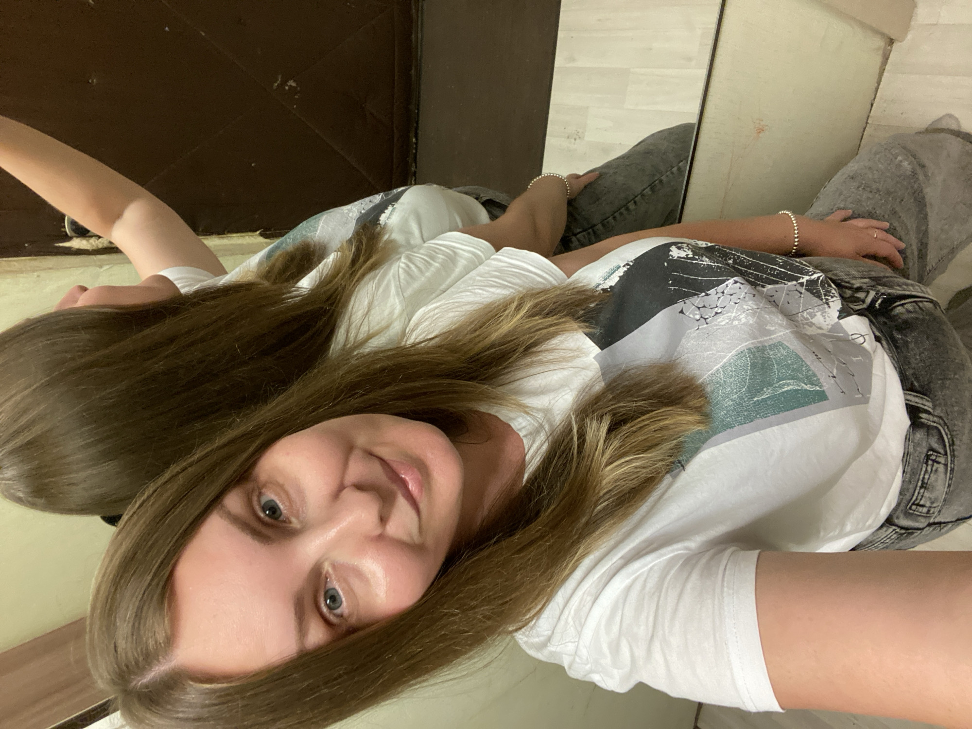 A fair-skinned woman with light brown hair and blue eyes is taking a selfie in a mirror. She is wearing a white t-shirt with a graphic print and gray jeans. The background includes a dark wooden door and a light-colored wall or paneling. The location is unknown, there is no mention of Noviy, Russia.