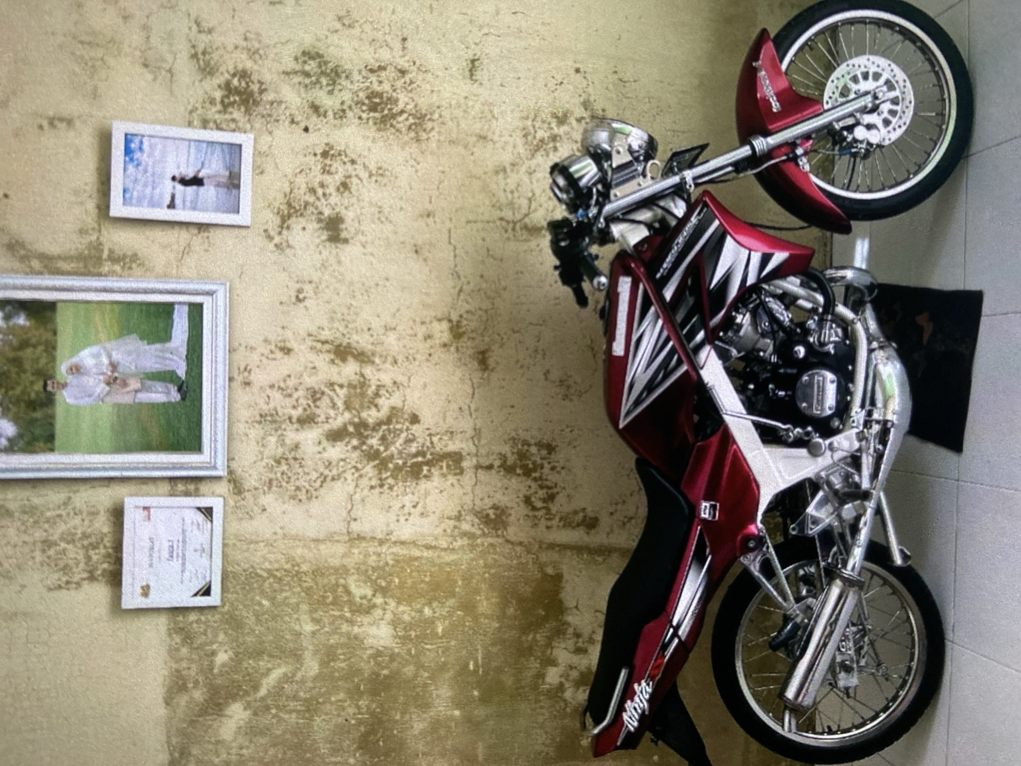 A red and silver motorcycle with "Ninja" written on its side leans against a mottled yellow wall. Three framed pictures hang on the wall to the left of the motorcycle. The top picture is rectangular and has a light-colored frame. The middle picture is larger, rectangular, and has a silver frame with a couple visible in the frame. The bottom picture is smaller, square, and has a white frame. The environment suggests an indoor location, possibly a garage or storage area. The location is unknown and cannot be determined as Lhokseumawe, Indonesia with the present image data.