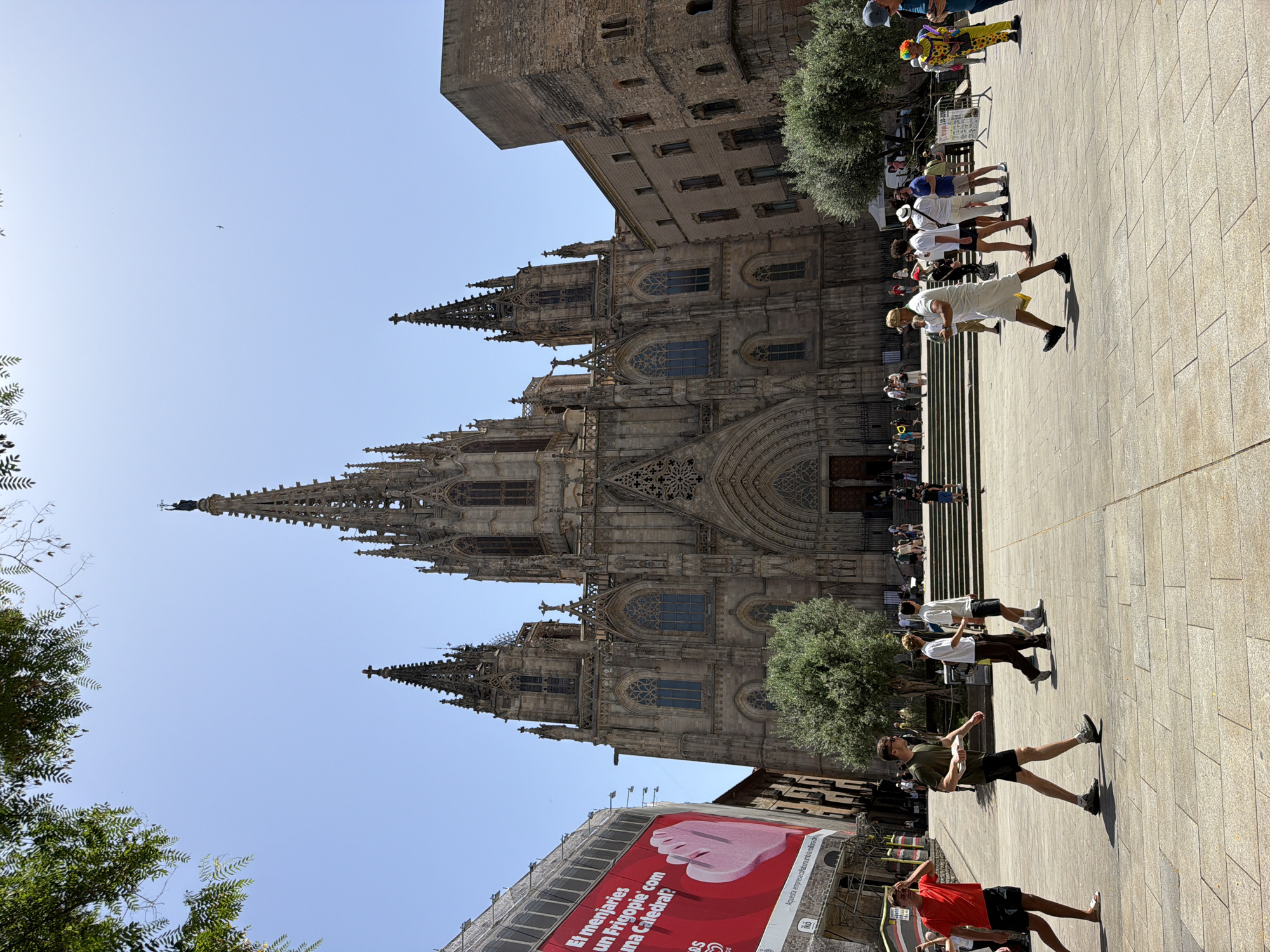 The image captures a view of the Barcelona Cathedral, a prominent Gothic Roman Catholic cathedral located in Barcelona, Spain. The cathedral's intricate facade, characterized by spires, arched windows, and detailed stonework, dominates the upper portion of the frame. In the foreground, a paved plaza is populated by various individuals.
Several people are walking across the plaza. On the left, a person in a red t-shirt and black shorts walks away from the viewer. Next to them, a person in a green t-shirt and black shorts, carrying a white object, walks towards the right. Further right, two individuals in casual attire, one in a white t-shirt and brown pants, the other in a white t-shirt and black shorts, are walking. A person in a white t-shirt and white shorts walks towards the left. To the far right, a person dressed as a clown in a yellow and black costume with colorful hair stands, and another person in a blue cap is partially visible.
In the background, to the left of the cathedral, a large banner with red and white graphics is displayed on a building under construction or renovation, with scaffolding visible. To the right of the cathedral, another building made of stone is visible. Green trees are interspersed throughout the scene, with some foliage in the upper left corner suggesting nearby trees or vegetation. The sky is clear and blue, indicating a sunny day. The overall scene depicts a bustling urban environment with tourists and locals interacting in the vicinity of a historic landmark.