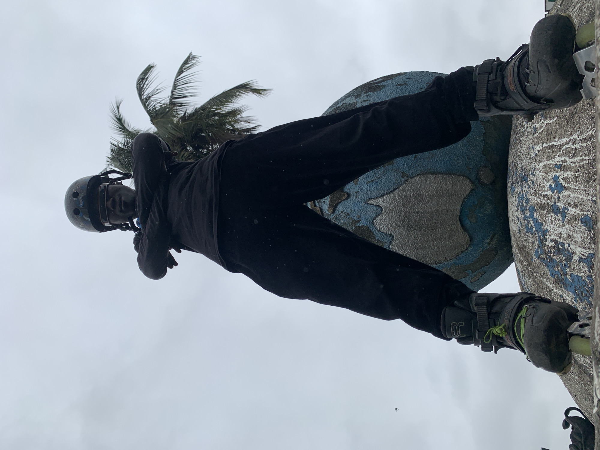 A low-angle, full-body shot shows a person wearing rollerblades, a helmet, and dark clothing, standing with arms crossed atop a weathered, globe-shaped sculpture. The sculpture is blue and grey with some peeling paint. The person's face is obscured by protective eyewear and the angle of the shot. A palm tree is visible in the background to the right, with a grey, overcast sky above. The image appears to have been taken outdoors. The scene is located in the city of Iganmu, Nigeria.