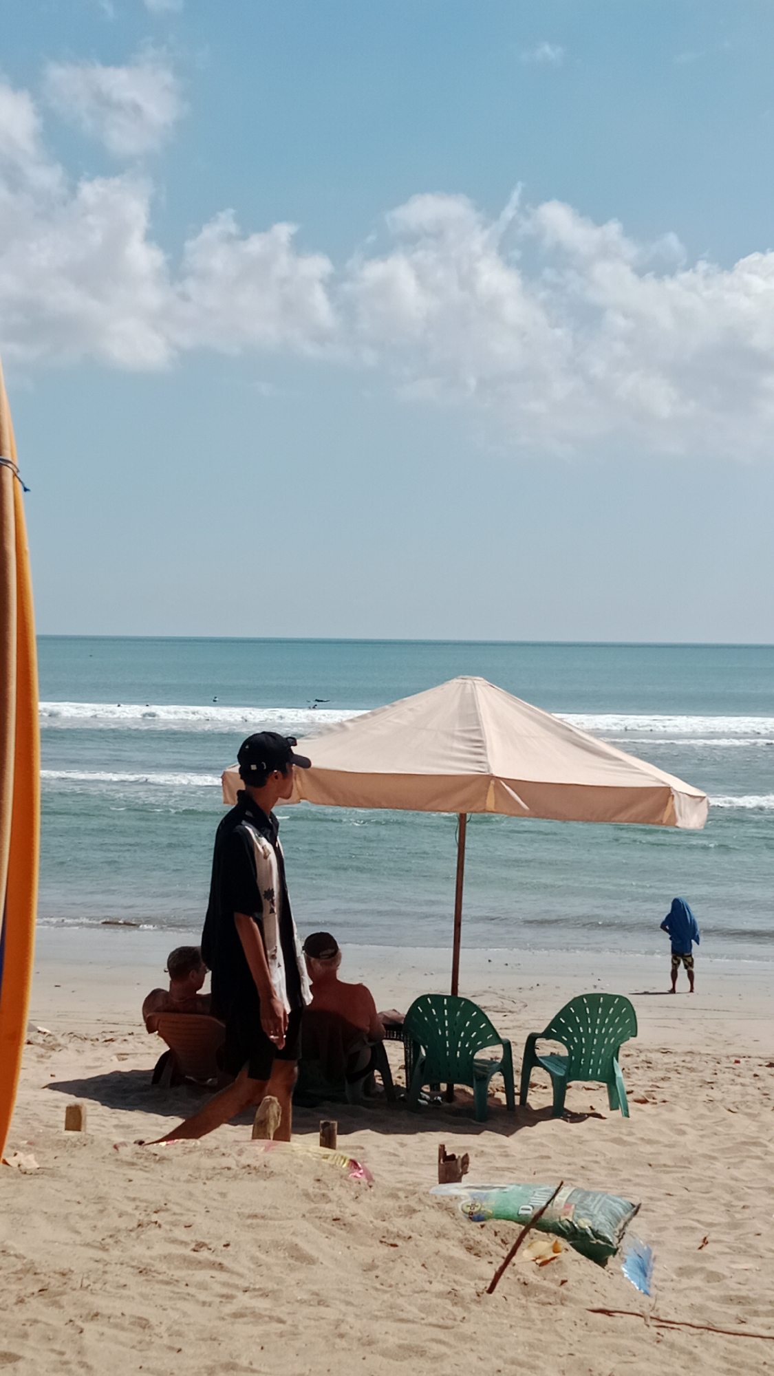 An outdoor, eye-level, vertical shot captures a sandy beach scene in Kuta, Indonesia, with the ocean stretching to the horizon under a bright, partly cloudy sky. In the foreground, a person wearing a black shirt with a white patterned collar and a black baseball cap is standing and facing right, looking out towards the ocean. Behind them, under a large, cream-colored umbrella, sit two green plastic chairs. To the left of the umbrella, three individuals are seated on lounge chairs, facing the ocean. One person, wearing a dark top and light bottoms, sits closest to the foreground. Further back, another person in a dark top is seated, and behind them, a person with lighter skin and dark hair is visible. In the middle ground, the ocean displays gentle waves. In the background, a person in a blue hooded garment stands with their back to the camera, facing the ocean, with several surfers visible in the water. On the left side of the frame, the edge of a yellow surfboard stands vertically. The sand is scattered with a few wooden posts and some trash.