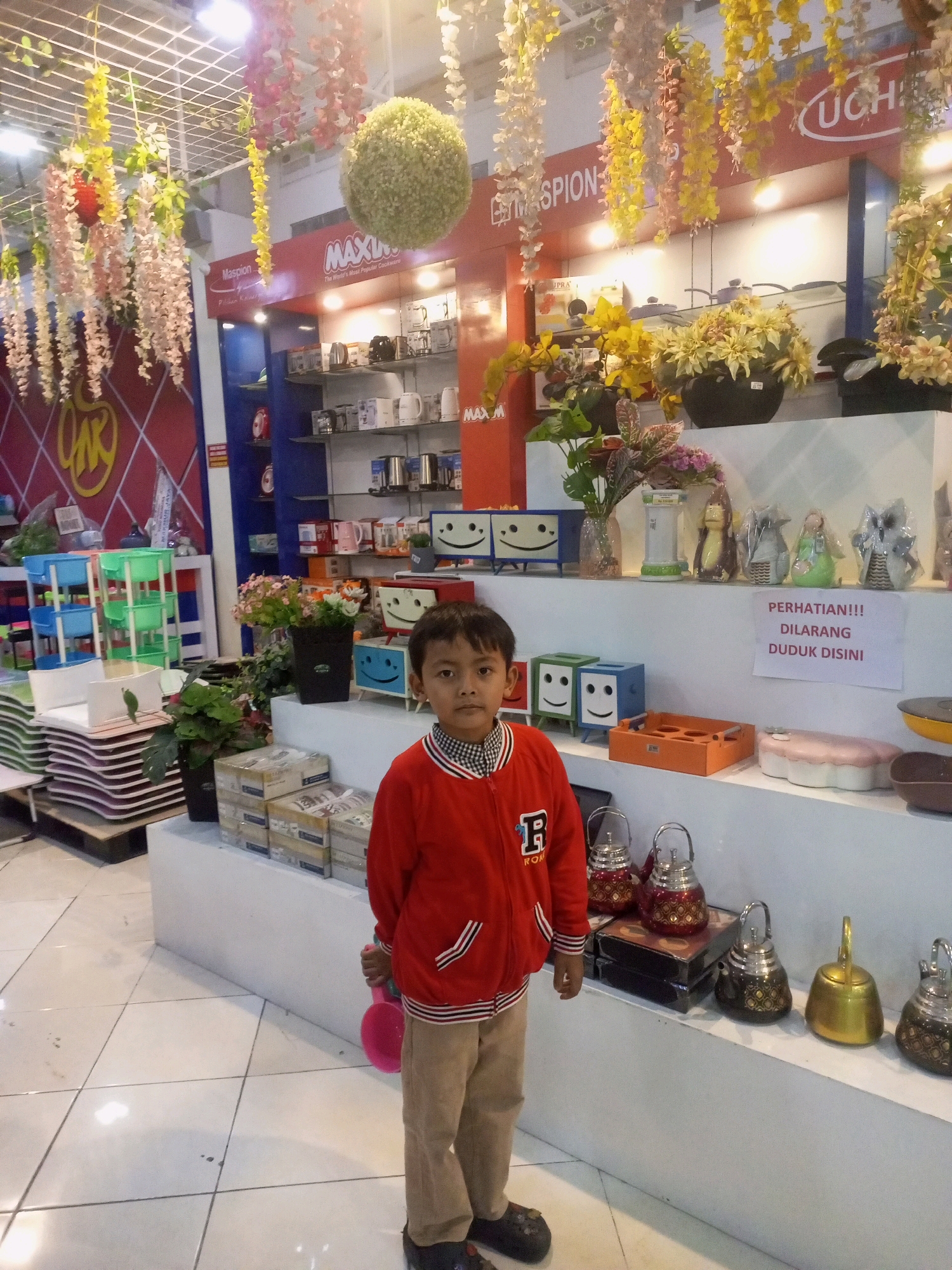 A young boy stands in a store, holding a pink shovel. He is wearing a red jacket with a white collar and beige pants. The boy is in the foreground of the image, looking towards the camera. Behind him, shelves are filled with various household items, including kitchenware, decorative objects, and plants. The store appears to be a department store or a homeware shop. Signs in the store are written in Indonesian. One sign reads "PERHATIAN!!! DILARANG DUDUK DISINI", which translates to "ATTENTION!!! NO SITTING HERE". The store has a bright, well-lit interior with white tiled floors. The store is located in the city of Ngasem, Indonesia.