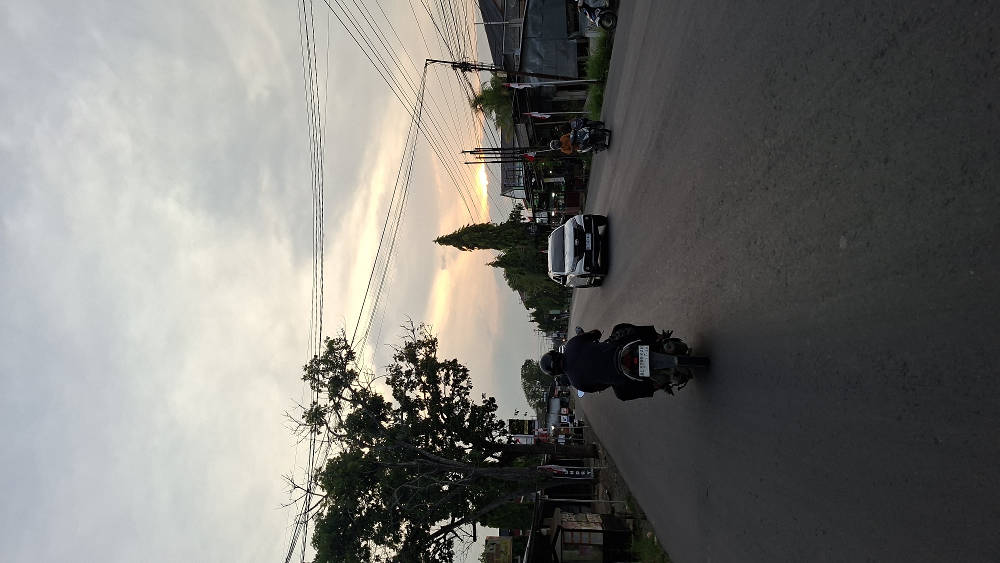 The image shows a street scene in Keudebayu, Indonesia, during what appears to be dusk. A person is riding a motorcycle in the foreground, seen from behind, facing away from the camera. The motorcycle has a license plate visible. In the middle ground, a white SUV is driving in the same direction. Further down the road, another person is riding a motorcycle. On the sides of the road, there are trees, buildings, and utility poles with numerous wires. The sky is cloudy with a warm glow from the setting sun visible on the horizon. The road is paved and appears to be a two-lane street.