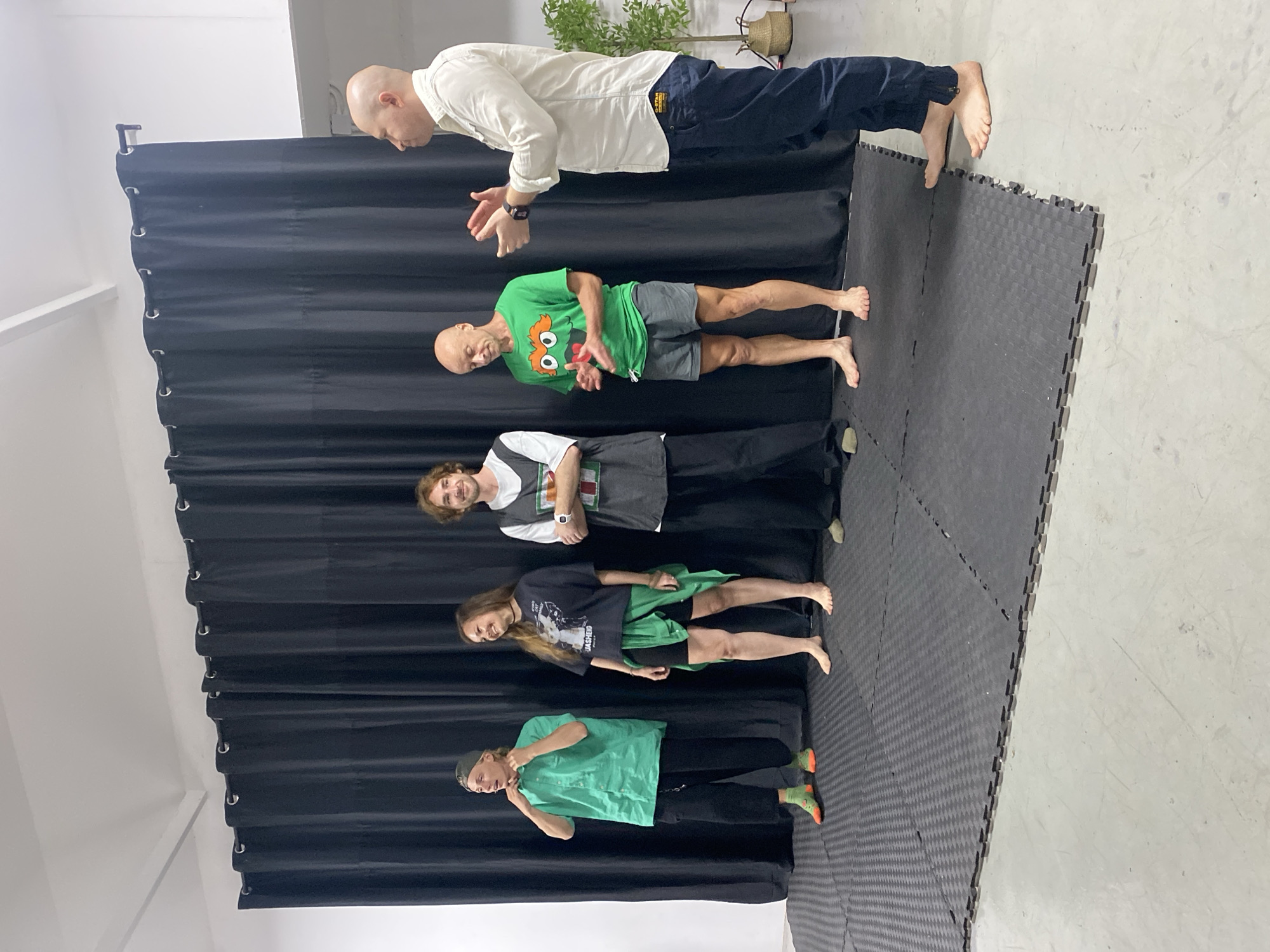 A group of five people are standing on a black mat in front of a black curtain. The person on the far left is wearing a green shirt and black pants, and has on orange and green socks. The second person from the left is a woman wearing a black t-shirt, black shorts, and a green sash. She is barefoot. The third person from the left is a man wearing a white and grey sleeveless shirt and black pants. The fourth person from the left is a man wearing a green t-shirt with a character graphic and grey shorts. He is barefoot. The person on the far right is wearing a white long-sleeved shirt and dark blue pants. He is clapping his hands. The environment appears to be an indoor studio space.