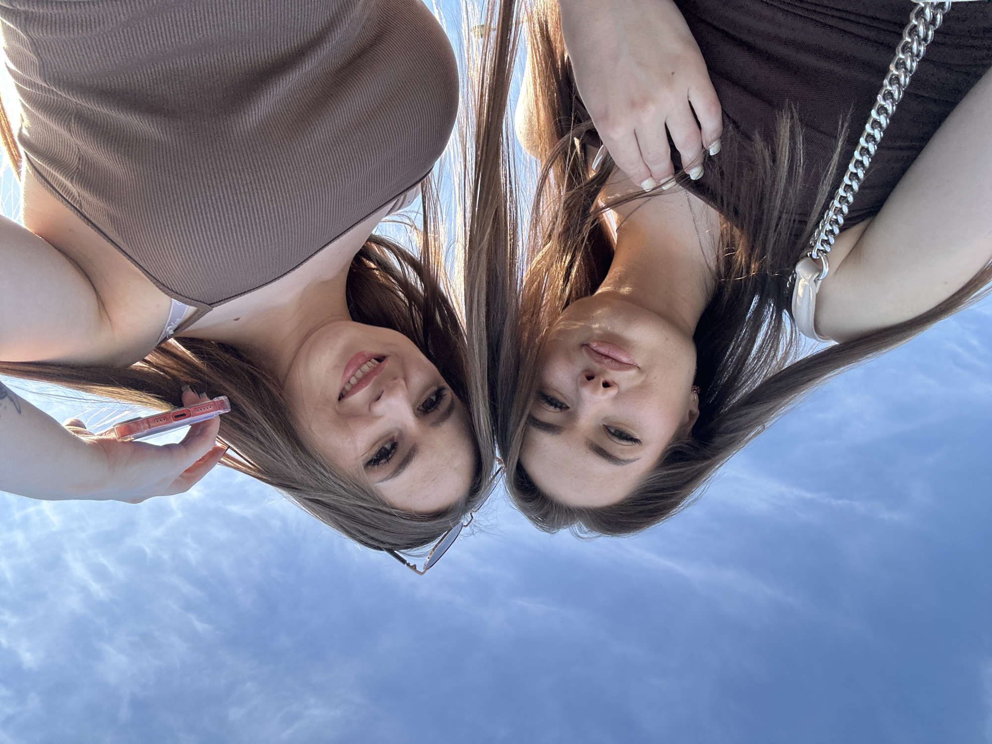 This image features two young women captured from a low-angle perspective, looking down towards the camera. The photo is oriented such that the clear blue sky forms the background, with the women positioned at the top of the frame. Both women have long, straight brown hair, which cascades downwards towards the sky.
The woman on the left is wearing a brown ribbed tank top or dress. She displays a cheerful smile, revealing her teeth. In her left hand, she holds a smartphone encased in a translucent pink or red case. A pair of sunglasses is visible near her chest, tucked into her attire. Her left arm is partially visible on the far left of the frame.
The woman on the right is wearing a dark brown ribbed garment, possibly a dress or top. Her expression is more neutral or a subtle, closed-mouth smile. A silver chain strap of a handbag is prominently visible, draped over her right shoulder and extending towards the center of the frame. Her left hand is near her face, featuring light-colored nail polish.
The background consists entirely of a bright, clear blue sky with sparse, wispy white clouds, indicating a daytime setting under natural sunlight. The strong lighting suggests the photo was taken outdoors. There is no discernible text visible in the image. The overall impression is one of casual friendliness between the two subjects.