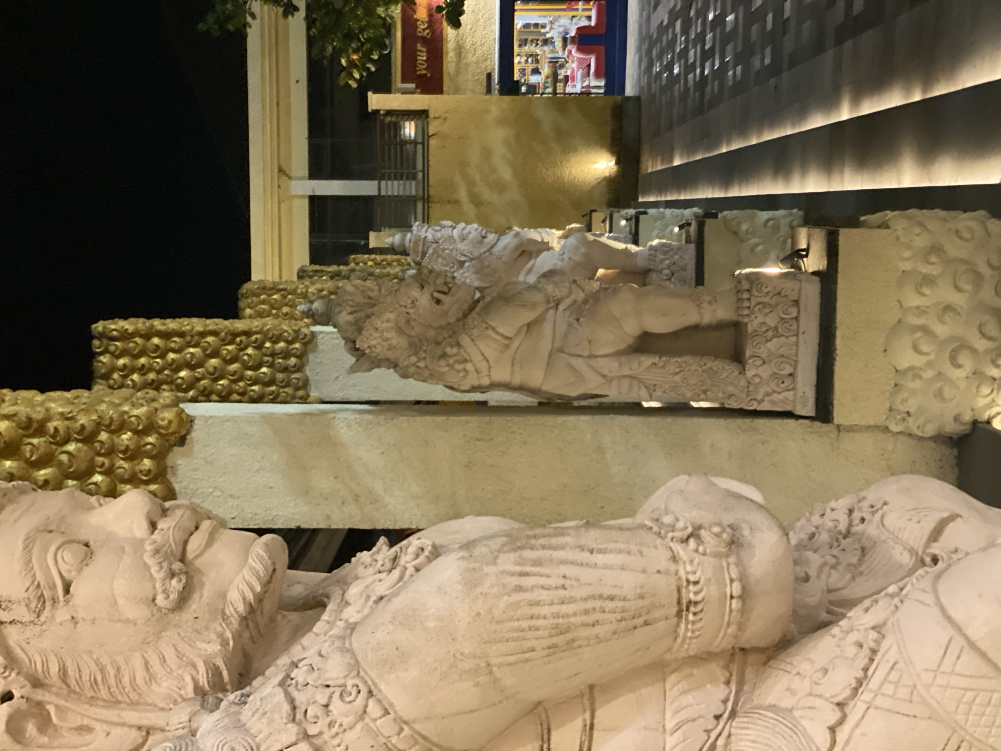An outdoor nighttime scene features a close-up, left-facing view of a weathered, light-colored stone statue with intricate carvings of a bearded face with a prominent mustache. The statue is positioned in the foreground, with its shoulder and chest visible. Behind it, a row of similar stone statues recedes into the background, each adorned with elaborate headdresses and intricate detailing. These statues stand against a pale yellow wall with ornate, gold-colored textured panels above. To the right, a building with a yellow facade is illuminated, revealing a red sign with white text that reads "your get". Further in the background, large windows show a brightly lit interior with red seating. A paved walkway with integrated strip lighting runs alongside the statues, leading towards the building. A tree branch with green leaves is visible on the far right, silhouetted against the dark night sky.