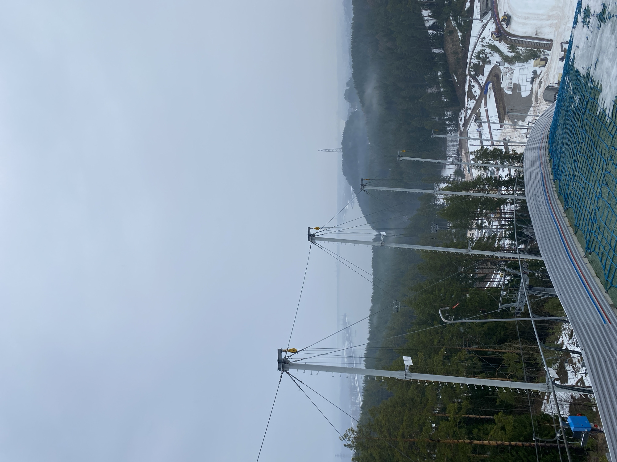 The image depicts the ski jumping hill "Снежинка" in Прикамский, Russia. The view is from the top of the inrun structure, looking down. The inrun is visible with its metal track and blue safety netting. In the background, there is a dense forest partially obscured by fog. Several ski lift support towers are visible extending through the trees. Snow is present on the ground in areas near the ski jumping hill. The sky is overcast, creating a muted, grey atmosphere.