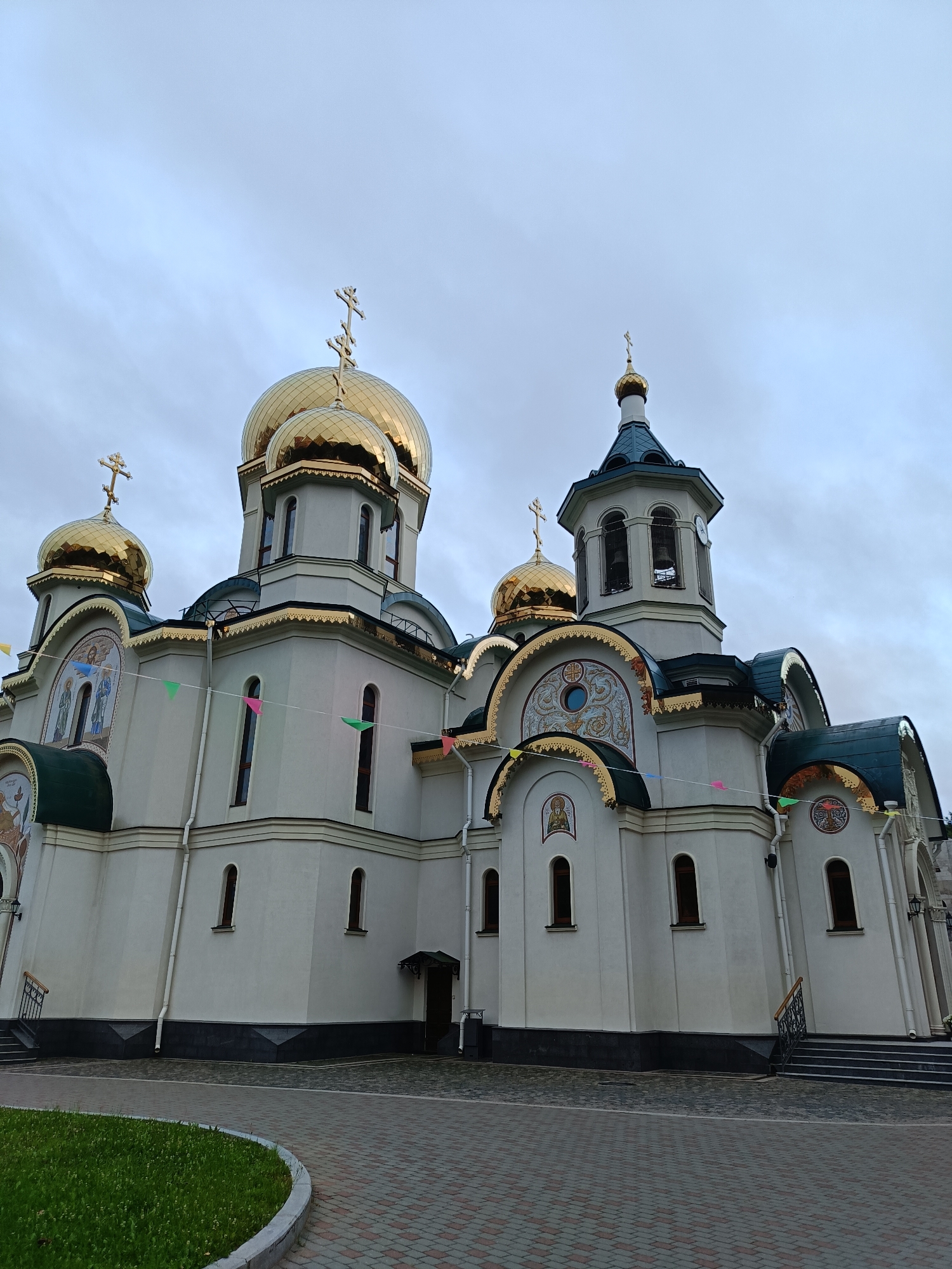 The image shows a large, cream-colored Orthodox church with multiple golden domes and a dark green roof. The church has intricate golden trim along its eaves and arches. There are several mosaic religious icons visible on the exterior walls, as well as arched windows. A bell tower with two bells is visible to the right of the main structure. The sky is overcast and gray. The foreground features a paved walkway and a curved patch of green grass. Small, colorful triangular flags are strung across the front of the building. No people are visible in the image. The architectural style suggests it is a Russian Orthodox church, possibly in Saint Petersburg, Russia, given the style.