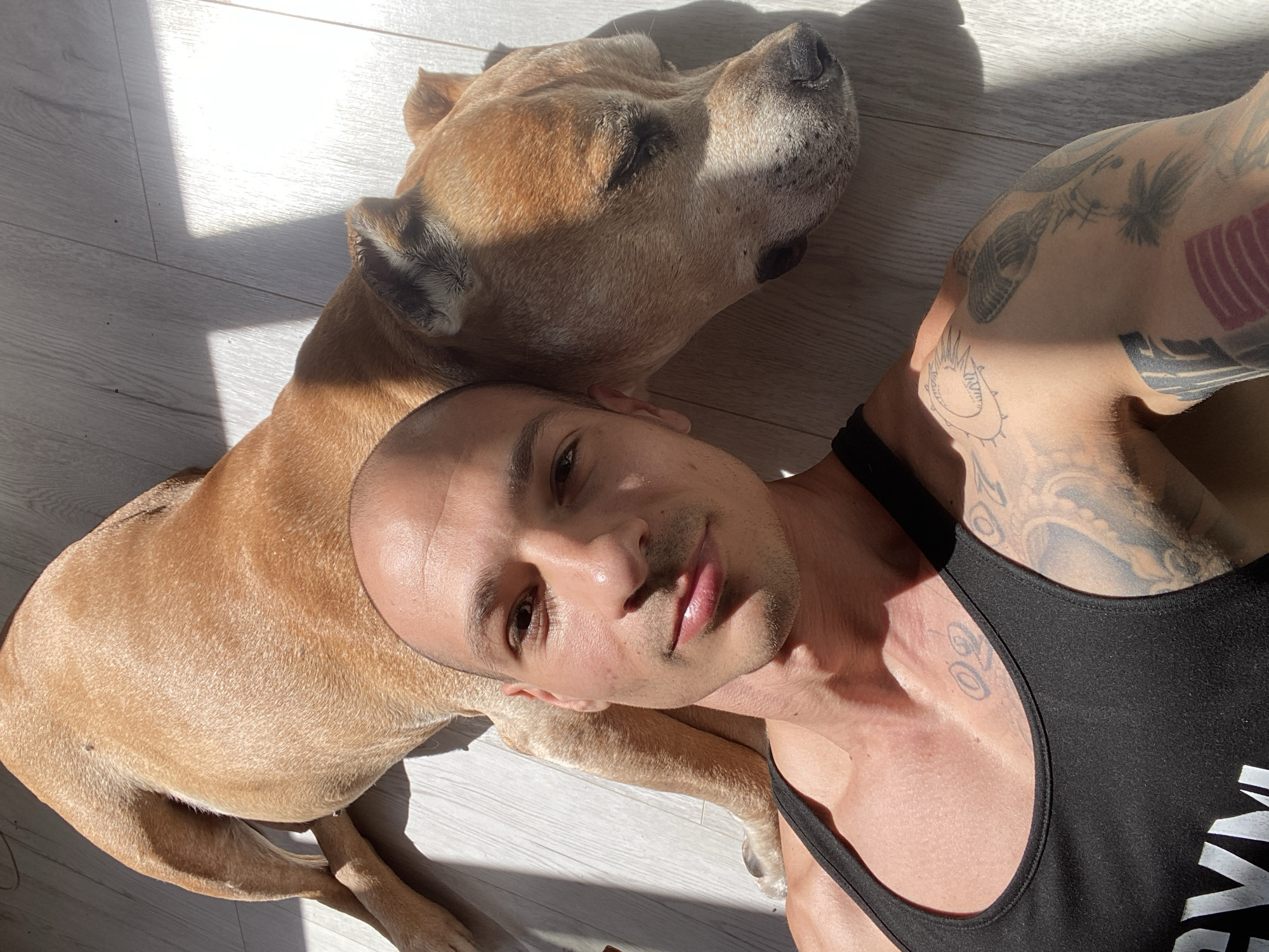 A man and a dog lie on a light-colored wooden floor. The man is bald, with brown eyes, and is wearing a black tank top. He has multiple tattoos on his arms and upper chest. The dog is light brown with short hair and is lying with its head near the man's head. Sunlight streams into the room, casting shadows across the floor. The location is unknown and cannot be determined from the image.