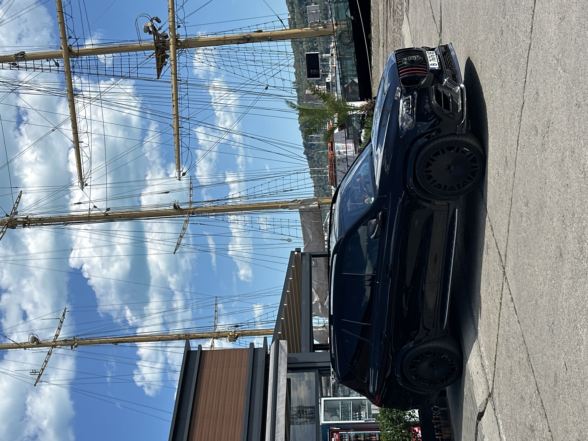 A black Brabus SUV is parked on a concrete surface next to a building with a wooden facade and glass windows. In the background, there's a tall ship with visible masts and rigging against a blue sky with white clouds. The location appears to be near the waterfront of Varna, Bulgaria, potentially near the Naval Museum, considering the presence of the sailing ship. The car has a license plate, "B 7676XB."