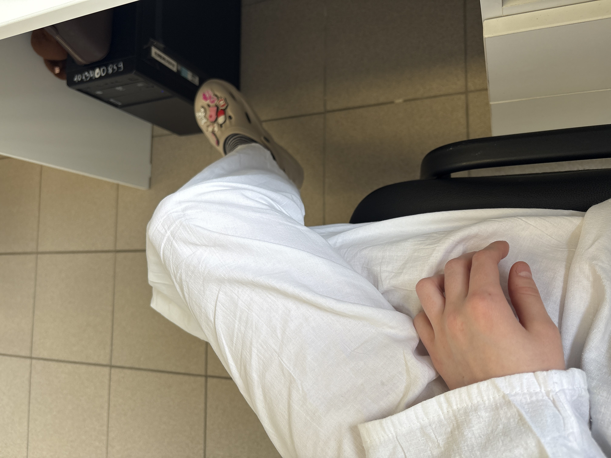 The image shows a person's legs and left hand. The person is wearing white pants and a beige clog-style shoe decorated with colorful charms. The background includes a tiled floor and a black computer tower. A dark chair arm is visible. The scene appears to be indoors, possibly an office or home environment. The lighting suggests daylight. There are no specific indicators of the location being in a city or country.