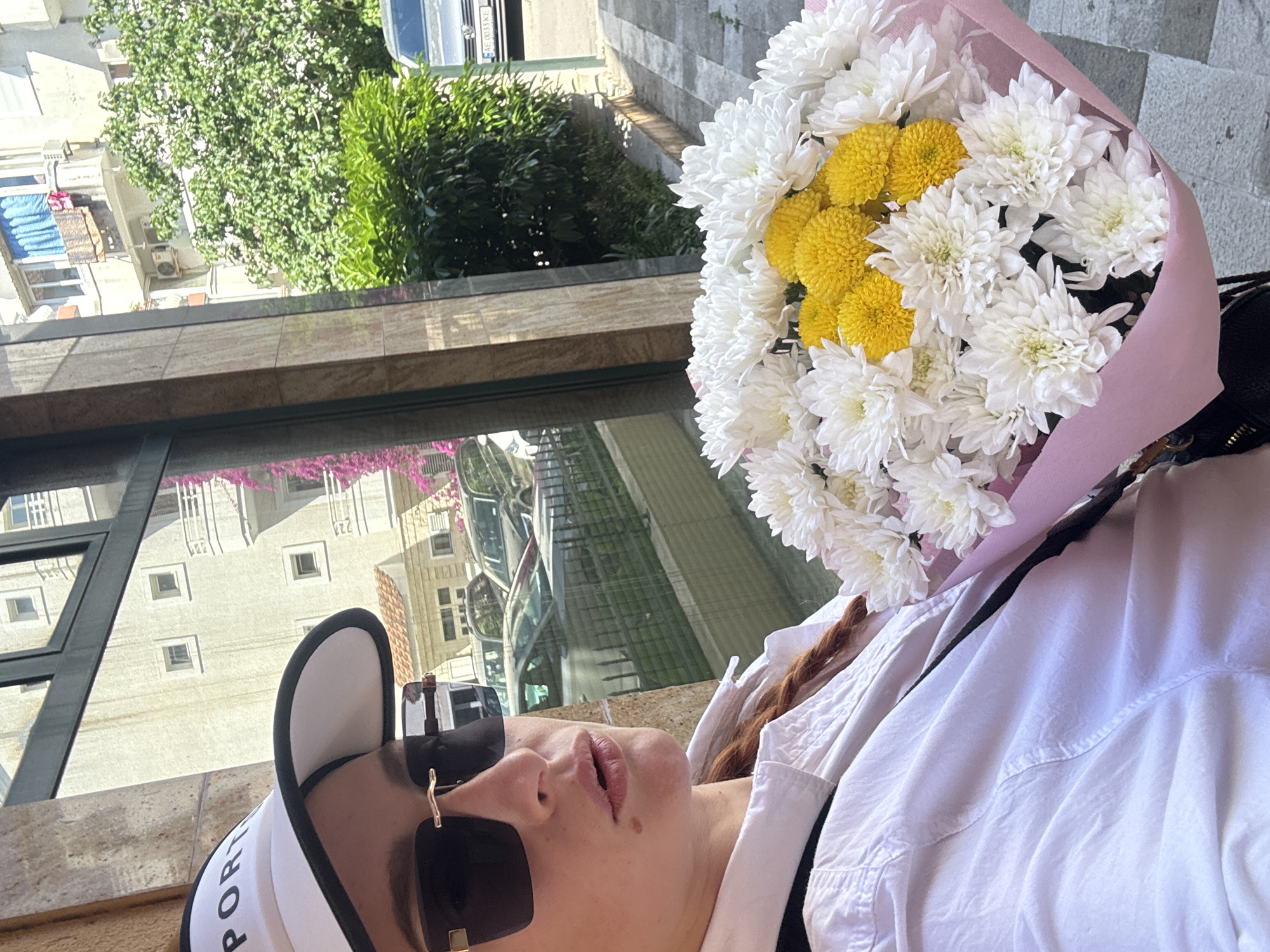 A low-angle, rotated shot depicts a woman holding a bouquet of white and yellow chrysanthemums wrapped in pink paper. The woman is wearing a white cap with the word "SPORT" partially visible, large sunglasses with gold accents, and a white button-down shirt.
The scene's background reveals a building facade with reflective windows that offer a distorted view of buildings, trees, and a parked car. The setting appears to be urban with architectural elements and greenery visible. Based on the reflected architecture, particularly the building with arched windows and terra cotta-colored roofs, it is consistent with the architectural style found in Budva, Montenegro. However, definitive location verification would require additional visual context or cross-referencing with local architectural features.