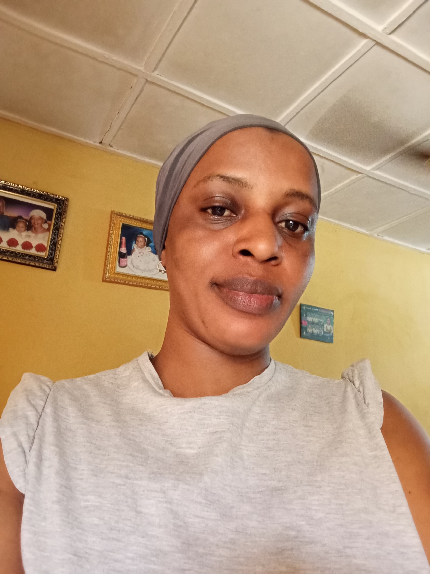 A medium indoor shot depicts a woman from the chest up. She wears a grey head wrap and a light grey t-shirt. Behind her is a yellow wall, adorned with two framed pictures and a small blue wall decoration. The ceiling is white with a grid pattern. The location is Narayi, Nigeria.