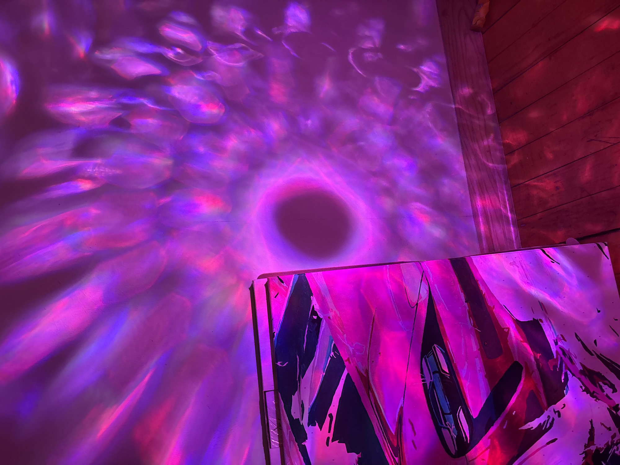 The image shows a section of a room with walls and a wooden corner. An abstract painting stands on the floor against the wall, illuminated in shades of pink and purple. The wall is covered in pink and purple light patterns with a dark circular shape near the center. The location is not discernible from the visual information provided. No people are present.