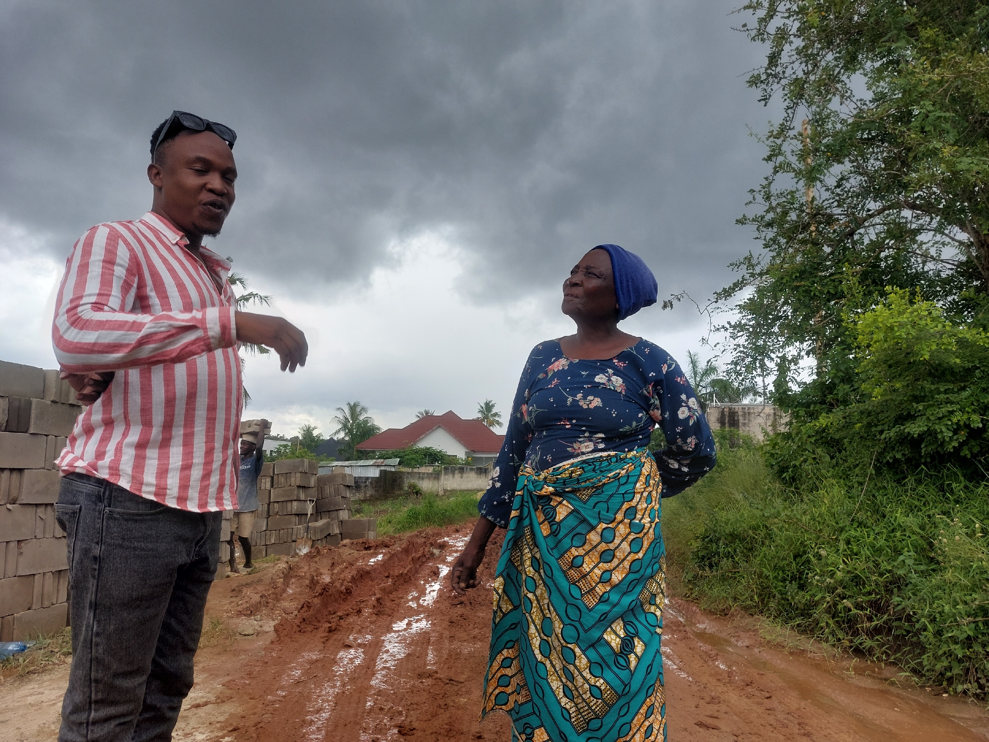 This outdoor photograph depicts two individuals, a man and an older woman, conversing on a muddy dirt road in what appears to be a developing area of Dar es-Salaam, Tanzania. The sky above is heavily overcast with dark gray clouds, suggesting either an impending storm or recent rainfall, which is consistent with the very muddy ground marked by vehicle tracks and puddles.
On the left, a man of East African descent stands, facing partially towards the woman. He wears a red and white striped long-sleeved collared shirt, dark jeans, and sunglasses perched on his head. His mouth is slightly open, and his hands are gestured, indicating he is speaking. To his left, large stacks of gray cinder blocks are visible, and in the mid-ground, another person is seen carrying blocks on their head, suggesting ongoing construction nearby.
On the right, an older woman of East African descent stands, looking upwards and to her right, away from the man. She wears a long-sleeved dark blue top with a floral pattern and a vibrant, patterned wrap skirt (likely a kanga or kitenge) featuring geometric designs in teal, gold, and dark brown/black. A plain blue headwrap covers her hair. Behind her, lush green vegetation and trees line the muddy road. In the distance, several houses with red roofs can be seen.
The scene conveys a sense of everyday life in a residential or semi-rural construction zone under a cloudy sky. No discernible text is visible in the image.