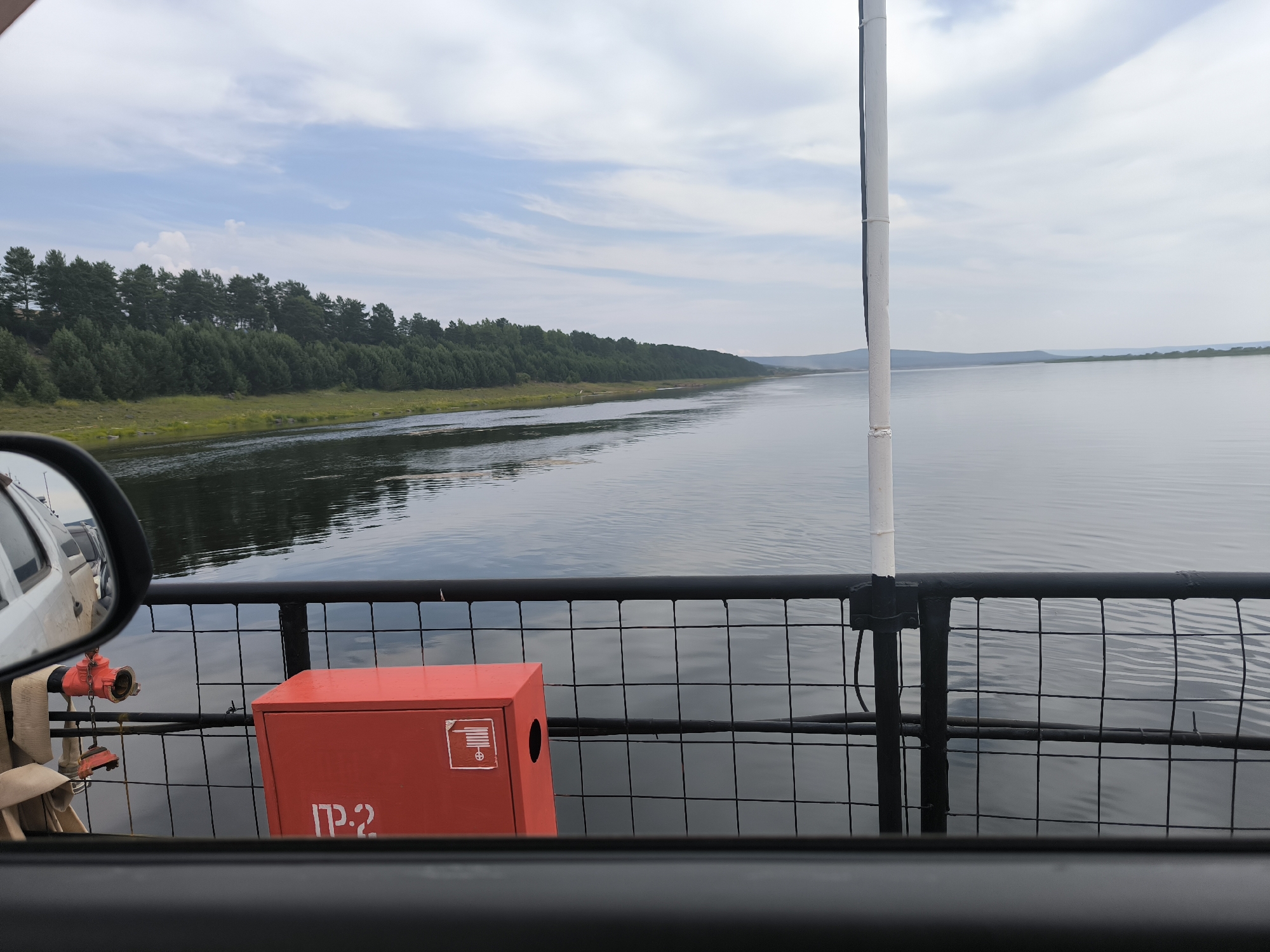 The image shows a view from a ferry crossing the Angara River in Boguchany, Russia. In the foreground, a red fire extinguisher box is visible on the deck of the ferry, secured by a black metal fence. The box has some Cyrillic lettering and a symbol on it. To the left, a portion of a white vehicle, likely a car, is reflected in a side mirror. The river stretches out to the horizon, with a lush green forest lining the opposite bank. The sky is partly cloudy, with a serene atmosphere. The ferry appears to be moving, as indicated by the gentle ripples on the water.