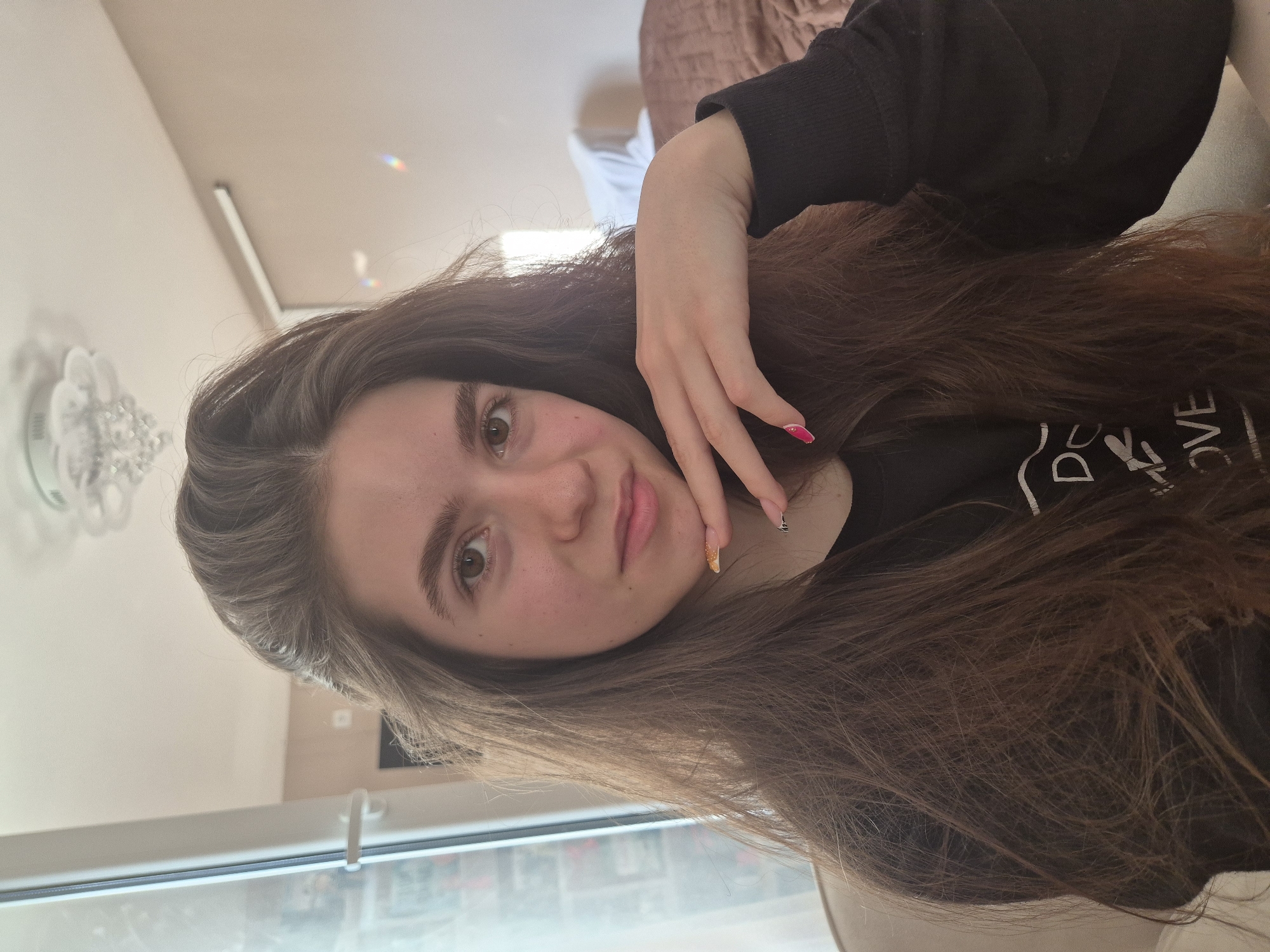A young woman with long brown hair poses for a selfie, tilting her head slightly. She wears a black shirt with the word "LOVE" printed on it. Her left hand rests on her chin, showcasing colorful nail polish. The background is a room with light-colored walls, a chandelier, a window, and a glimpse of the outdoors. There is no information to indicate this is Pavlikeni, Bulgaria.