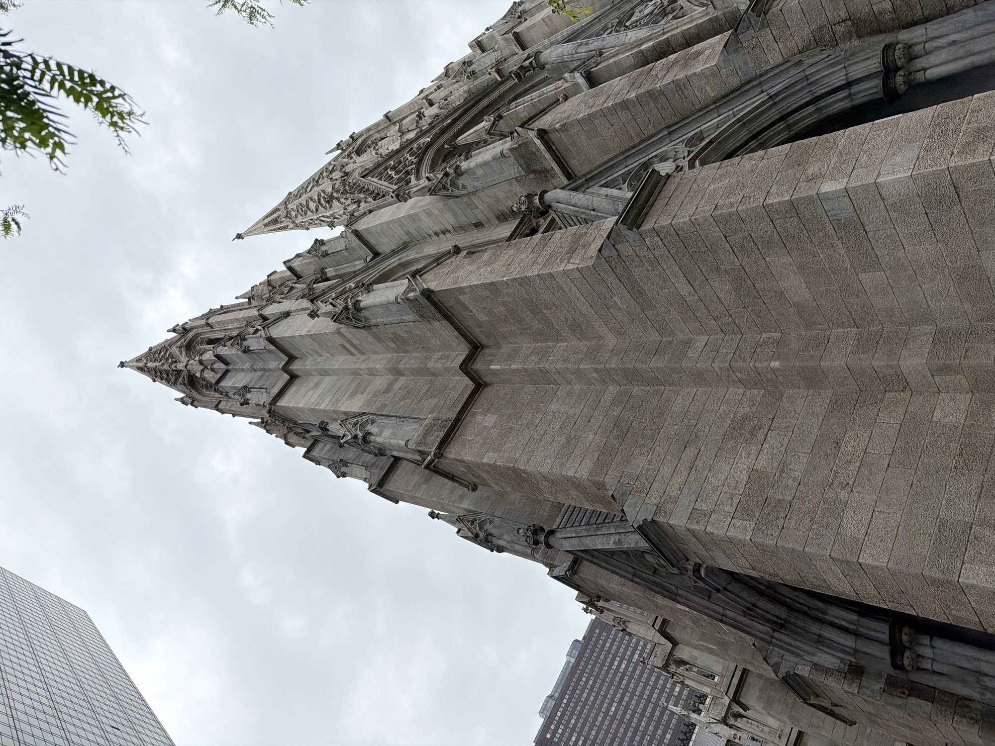 The image presents a low-angle perspective of a gothic-style cathedral, identified as St. Patrick's Cathedral in New York City, United States of America. The cathedral's facade is predominantly composed of gray stone blocks, exhibiting a rough, textured surface. The architectural details include pointed arches, spires, and vertical ribs, typical of the Gothic Revival style. The sky is overcast. The architectural details of a neighboring modern building can be seen. A few small branches are visible in the upper left.