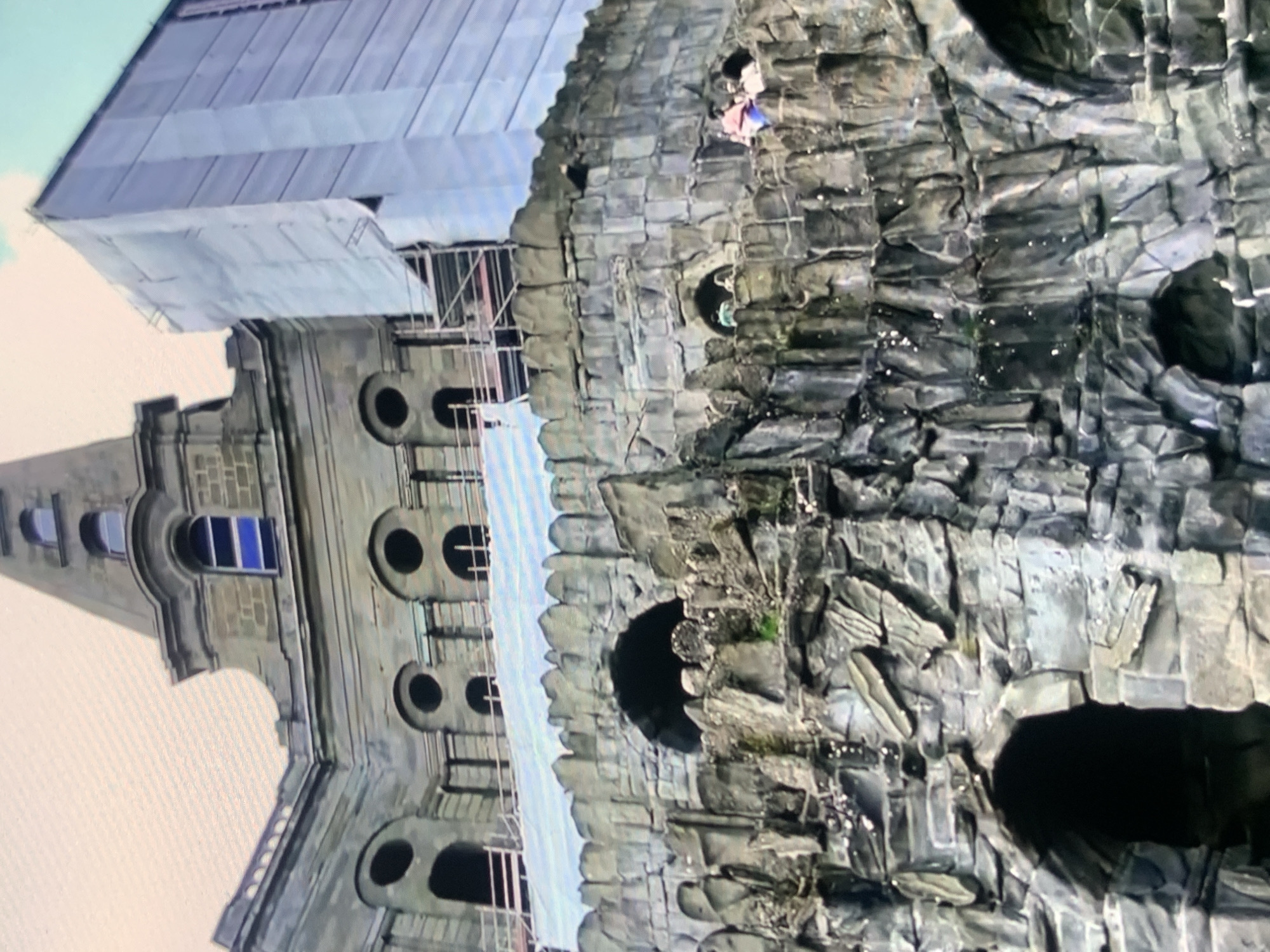 The image shows the exterior of a building resembling a medieval castle, with architectural elements such as turrets, arched windows, and stone detailing. Part of the building is covered in scaffolding and a white protective sheet, indicating ongoing construction or renovation. In the foreground, there is a rocky, artificial landscape with arches and openings, also appearing to be part of the structure. Several people are visible on a ledge of the rocky structure, some of whom appear to be interacting. The sky is visible in the upper portion of the image. The location in Stavropol, Russia, is not discernible from the visual information presented in the image.