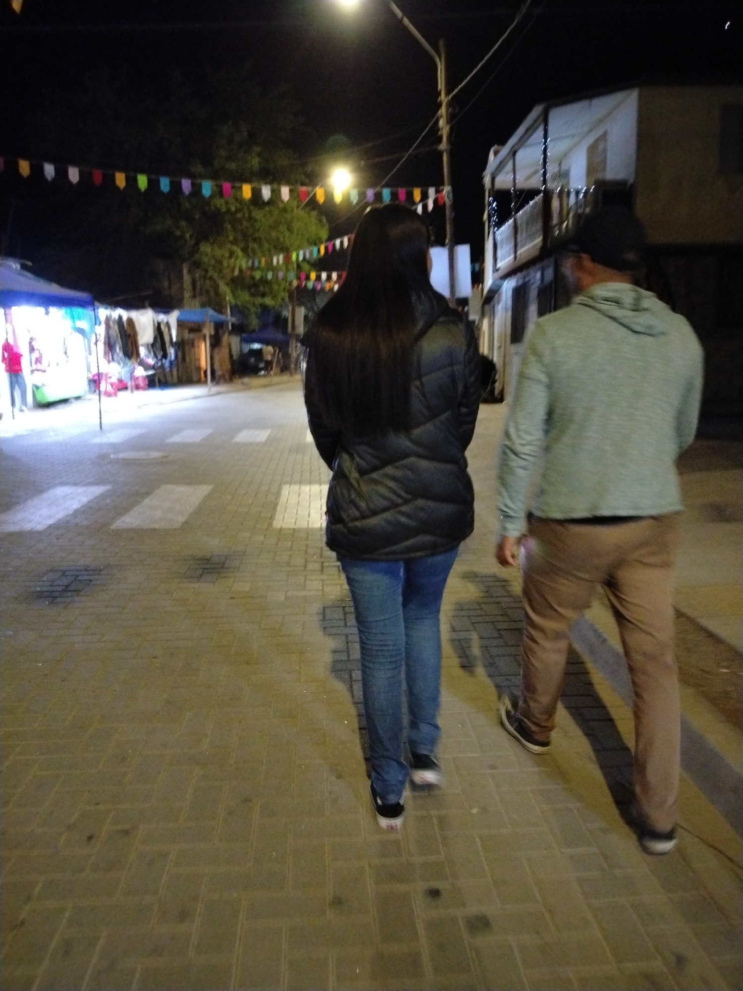 Two people are walking away from the camera on a cobblestone street at night. The person on the left is wearing a black puffy jacket and blue jeans. The person on the right is wearing a light green hoodie and brown pants. There are colorful banners strung across the street in the background, and a building with balconies is visible on the right side of the image. The scene appears to be a street market or festival in Pozo Almonte, Chile.