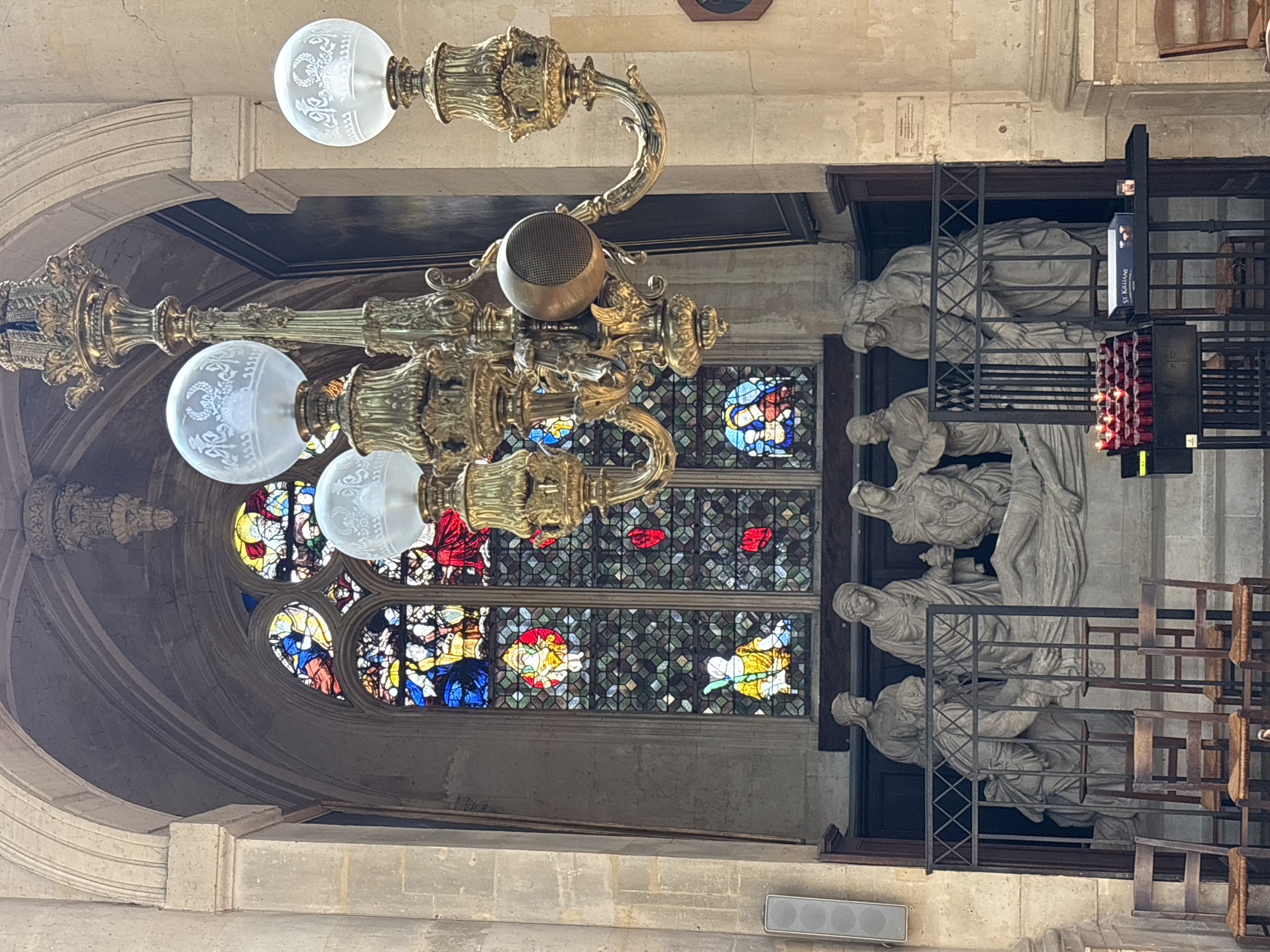 This is an interior shot of the Saint-Séverin Church in Paris, France, capturing a religious scene within a Gothic architectural setting. The focal point is a large, ornate gilded chandelier hanging prominently in the foreground, featuring multiple glass globes with intricate etched designs. Behind the chandelier, a large stained-glass window displays vibrant religious imagery in geometric and figural patterns. Below the window is a stone sculpture depicting the deposition of Christ, with several figures gathered around a central figure. The sculpture is enclosed by a metal railing. In front of the sculpture, a rack of lit red votive candles is arranged on a black stand, alongside a box labeled "ST. KILLIANS." Several wooden chairs with woven seats are positioned in the lower left portion of the frame, facing the sculpture. The stone walls and vaulted ceiling of the church are visible, contributing to the solemn and historical ambiance.