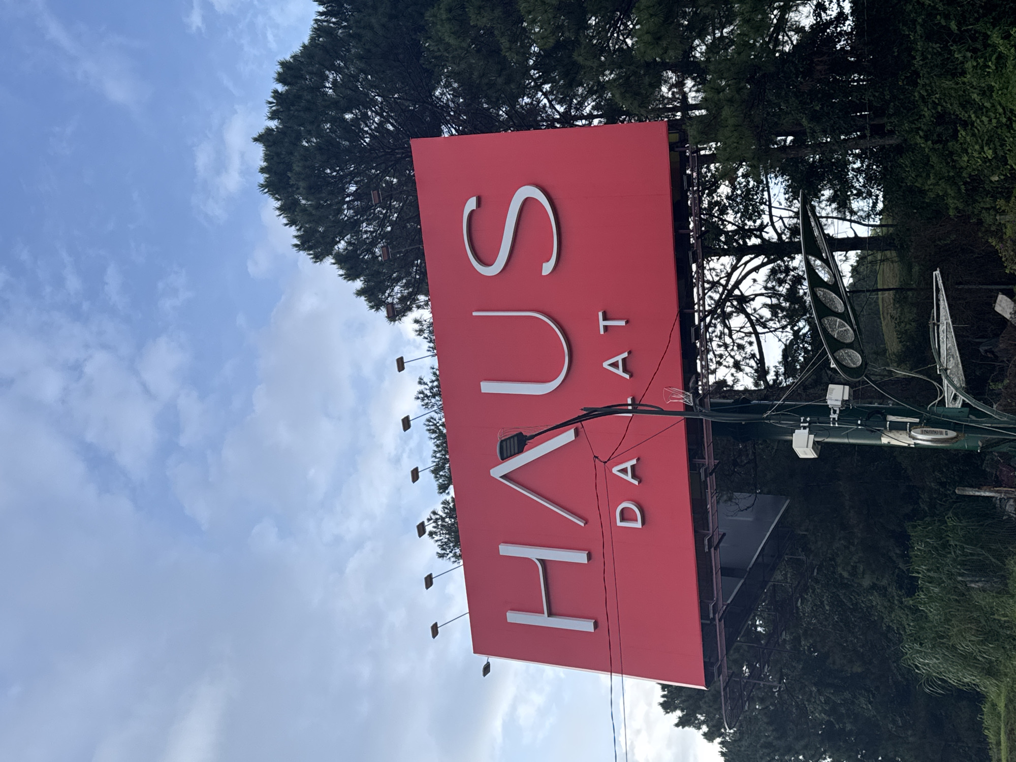 A bright red billboard in Da Lat, Vietnam, displays the word "HAUS" in large, white, stylized capital letters. Below "HAUS", the word "DALAT" is written in smaller, white capital letters. The billboard is supported by a dark metal frame and a green pole that runs vertically in front of it. Several wires and electrical components are attached to the pole. Above the billboard, a dense canopy of dark green pine trees is visible against a cloudy blue sky.
