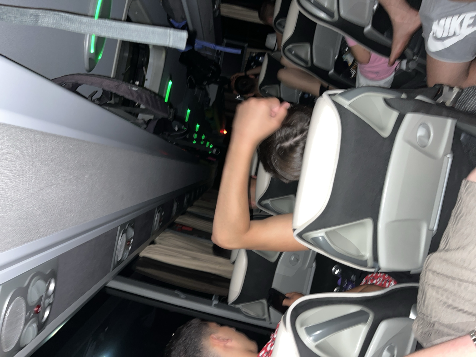 The image depicts the interior of a bus. People are seated. A person wearing Nike shorts is visible. Seats are upholstered in black and white. Some overhead compartments and lights are visible. The image location is Popovitsa, Bulgaria.