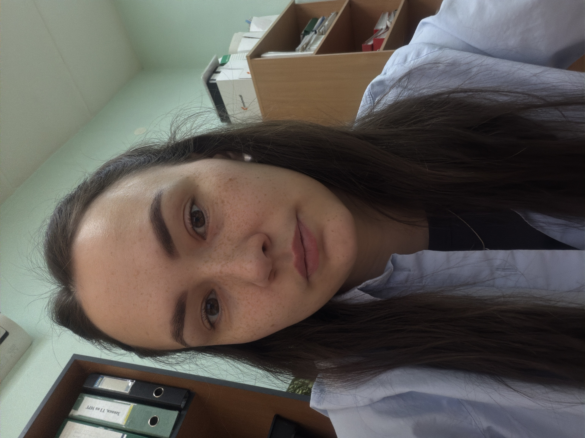 A young woman with brown hair and freckles is looking at the camera. She is wearing a light blue shirt. In the background, there is a wooden shelf with various items and a shelf with folders labeled in Russian. The location is potentially an office or administrative space in Chaikovsky, Russia, based on the labeling of the folders.