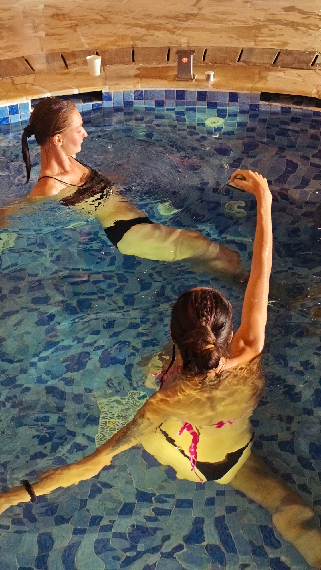 The image captures two women relaxing in a pool or jacuzzi, likely indoors or during the evening, characterized by warm, artificial lighting. The water is clear, revealing the blue and light blue mosaic-patterned tiles lining the pool floor.
In the foreground, a woman is seen from behind, her dark hair tied in a bun with a yellow elastic. She wears a yellow bikini top with pink straps and black bottoms, and tattoos are visible on her left arm. Her right arm is raised, holding a small, light-colored object, possibly a waterproof gadget or a sponge.
Further back and to the left, another woman is partially submerged, viewed in profile. Her hair is pulled back into a ponytail, and she is wearing a dark-colored swimsuit, possibly black. Her expression appears calm as she looks towards the left.
The rim of the pool is made of light brown, stone-like tiles. On this edge, a white disposable cup, a dark rectangular fixture with a red light, and a small metallic, cylindrical object are visible. The scene conveys a sense of relaxation and leisure. There is no visible text in the image.