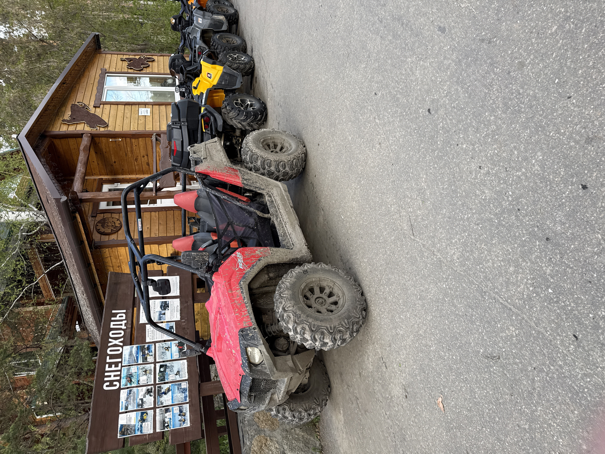 The image shows a row of four-wheeled all-terrain vehicles (ATVs) parked on a paved surface next to a wooden building and a sign in Russian, suggesting a rental or tour operation. The first ATV is red and covered in mud, indicating recent use. Behind it are other ATVs in varying conditions, including yellow and black models. The wooden building features decorative wooden cutouts, and the sign displays pictures and information about "снегoxoды", likely snowmobiles or similar winter equipment, suggesting the location caters to off-road adventures in different seasons. The location is likely Zolotoi Plyazh, Russia, due to the Russian signage and architectural style. The scene appears to be set during a spring or autumn season, as there are trees visible with green leaves.