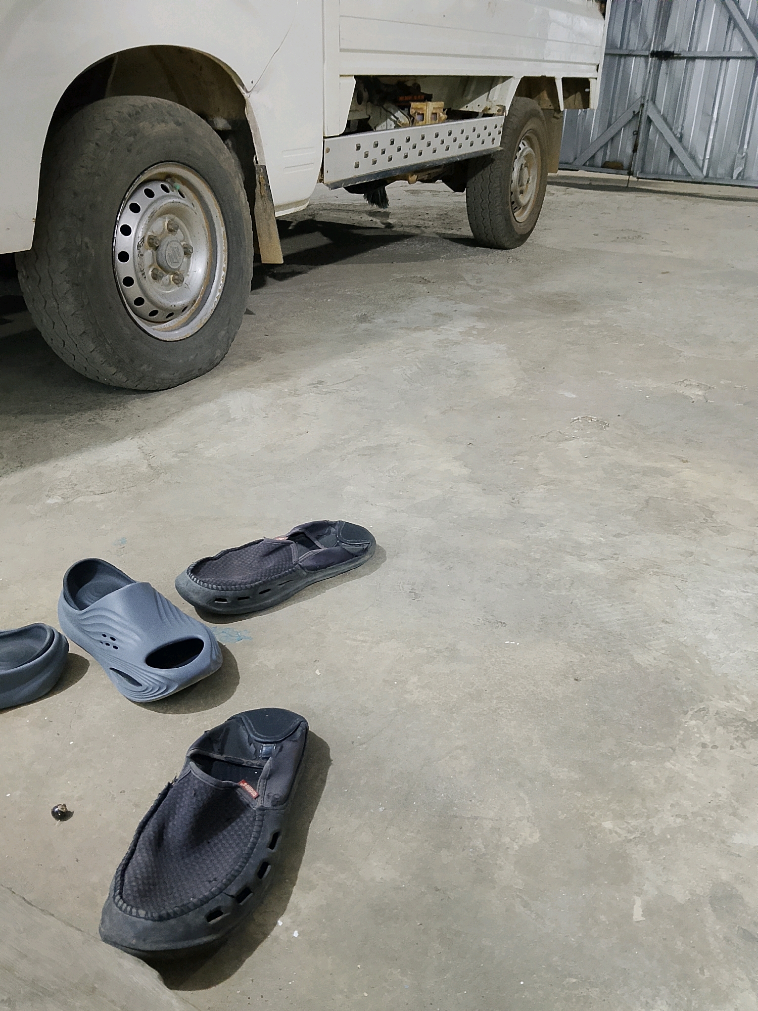 This is a medium-angle, eye-level shot of a white utility truck parked on a concrete floor, likely in a garage or workshop in Negeri Agung, Indonesia. Three pairs of shoes are scattered on the floor in the foreground. Two dark-colored slip-on shoes are on the right, one with its opening facing left, and another slightly further to the left, open to the right. A pair of gray slides with a rippled design are on the left, with one of them partially visible from the side. A small, dark, spherical object, possibly a bolt or a bead, rests on the concrete between the gray slides and the dark slip-on shoes. The truck's front passenger-side tire and wheel are visible on the left, and the truck's undercarriage, including a metal step, is visible extending from the passenger side. The background features a corrugated metal wall. The lighting appears to be artificial and casts shadows on the floor.