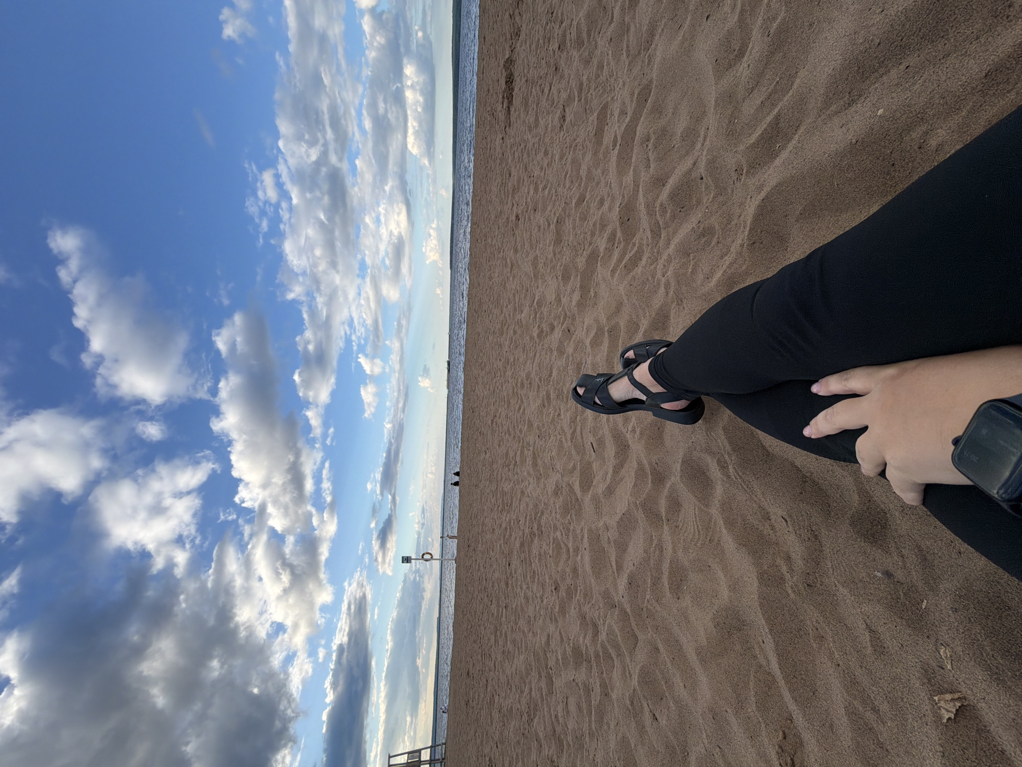This media file depicts a person's legs and feet resting on a sandy beach. The person is wearing black pants and black sandals. They are also wearing a dark-colored smartwatch on their left wrist. The scene shows a wide expanse of sand with ripples indicating the texture of the beach. In the background, the calm sea meets a sky filled with dramatic clouds. The sky transitions from a bright blue near the horizon to darker hues higher up. The orientation of the image suggests it was taken with the camera pointed towards the sky and horizon, with the person's legs and feet in the foreground. The overall impression is one of relaxation and enjoying a peaceful moment by the water. The lighting suggests it could be late afternoon or early evening, with soft, diffused light illuminating the scene. The presence of a life preserver on a pole in the distance suggests this is a public beach.