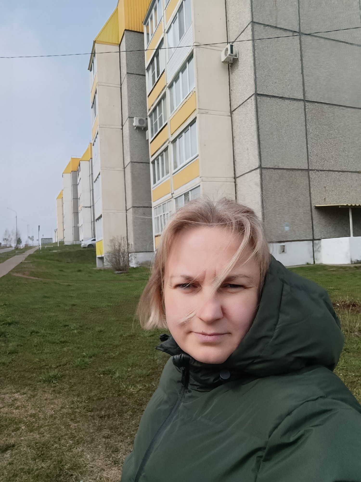 A blonde woman takes a selfie outdoors on what appears to be an overcast day in Марковский, Russia. She is in the foreground, wearing a dark green puffy jacket with a hood, which is pulled up. Her hair is slightly windblown, and she has a serious or neutral expression.
Behind her, a grassy slope leads up to a paved path or road. In the midground and background, a row of multi-story residential buildings with light-colored facades, grey concrete panels, and distinctive bright yellow roofs line the landscape. The architecture suggests a typical post-Soviet apartment block style. White-framed windows are visible on the buildings, and at least one outdoor air conditioning unit is attached to a wall. Power lines can be seen stretching across the light grey sky above the buildings. The ground is covered in green grass, indicating a temperate climate. No specific activity beyond taking the photo is apparent. There is no visible text.
